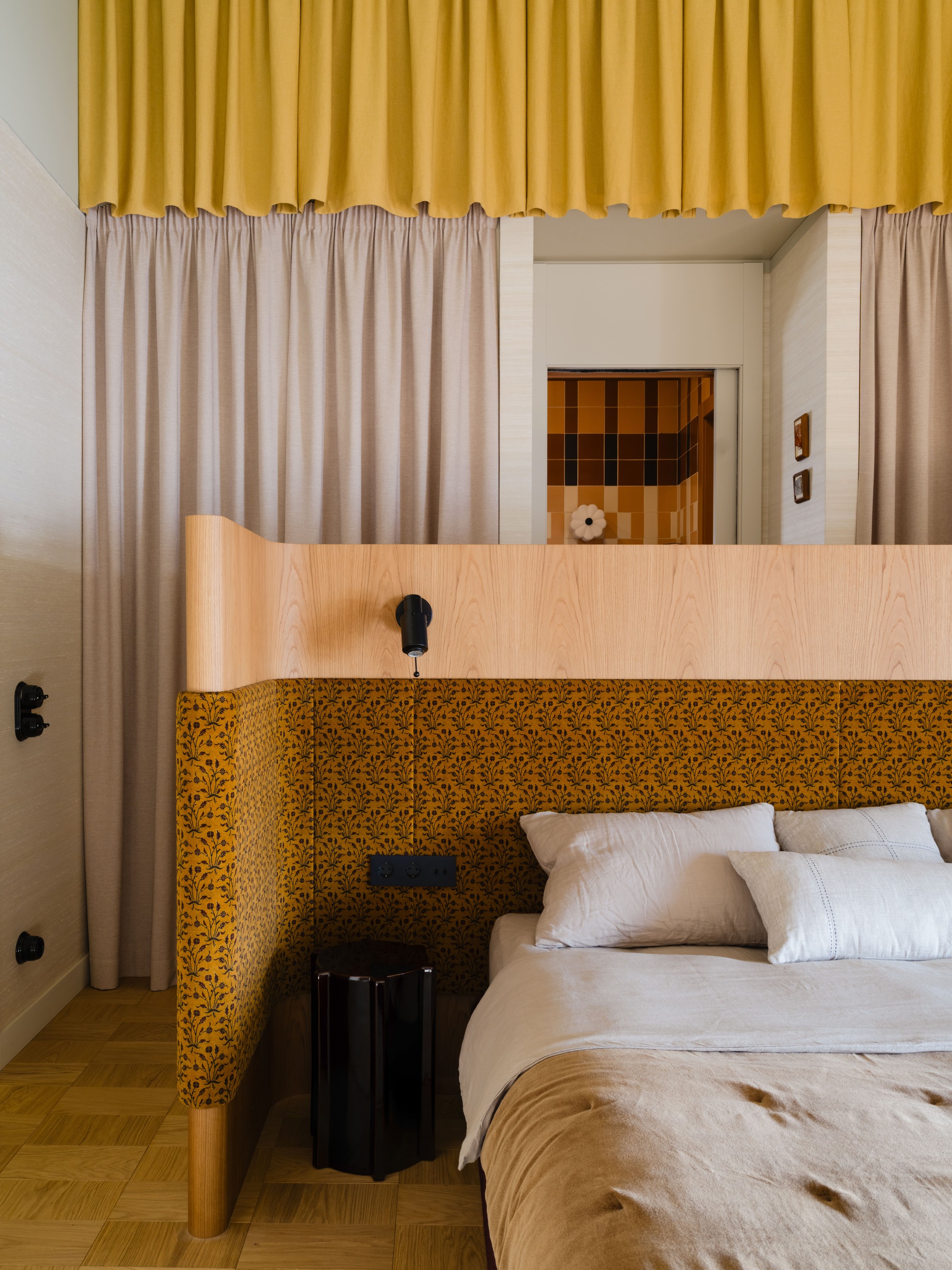 Bed with curved wooden headboard upholstered in mustard floral fabric, beneath gathered curtains and a view into a tiled bathroom