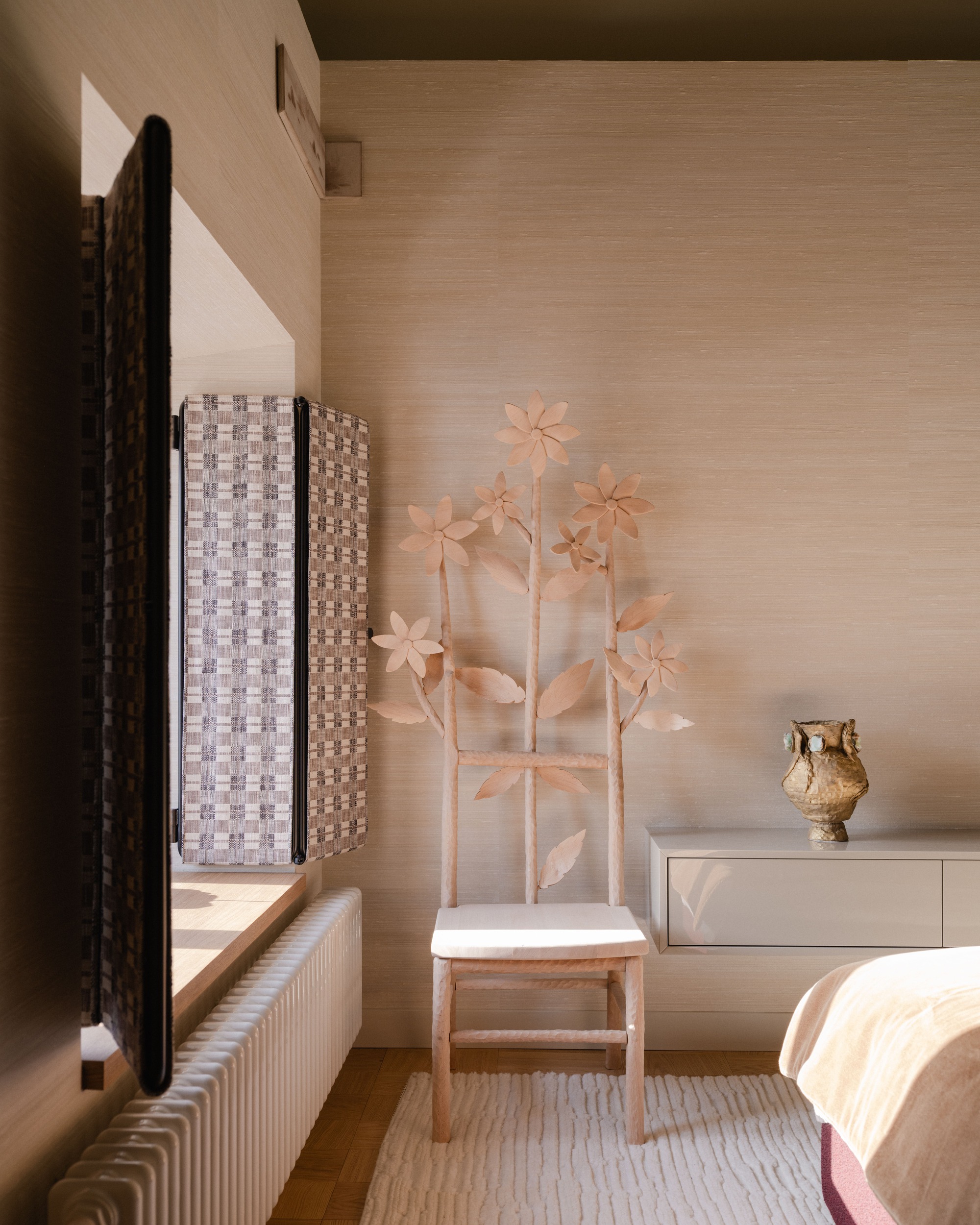 Carved wooden chair with flower-shaped leaves beside a window with woven shutters, a radiator and a ceramic vase in warm light