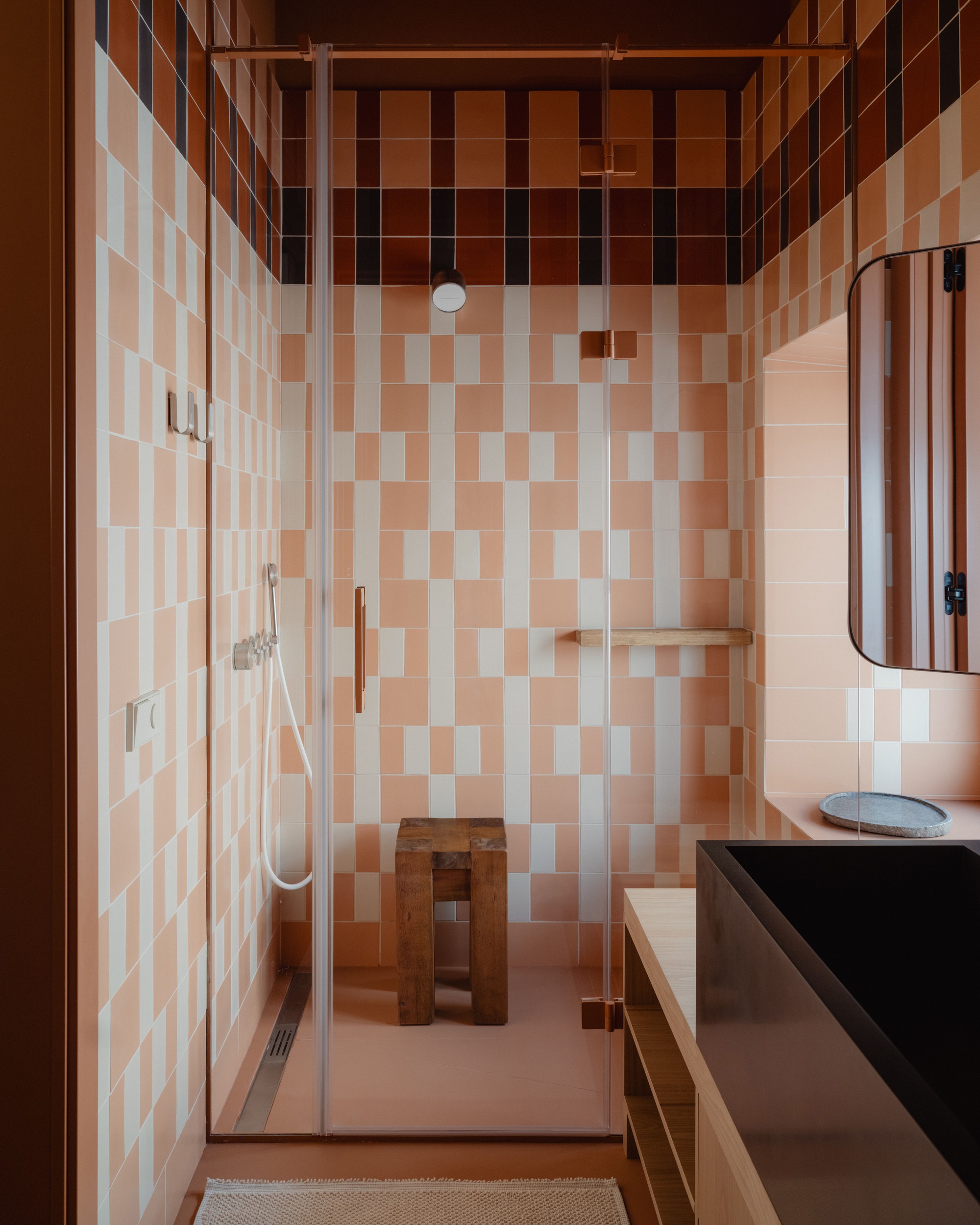 Shower enclosure lined in pink and white rectangular tiles with a dark red band, a wooden stool and a black basin