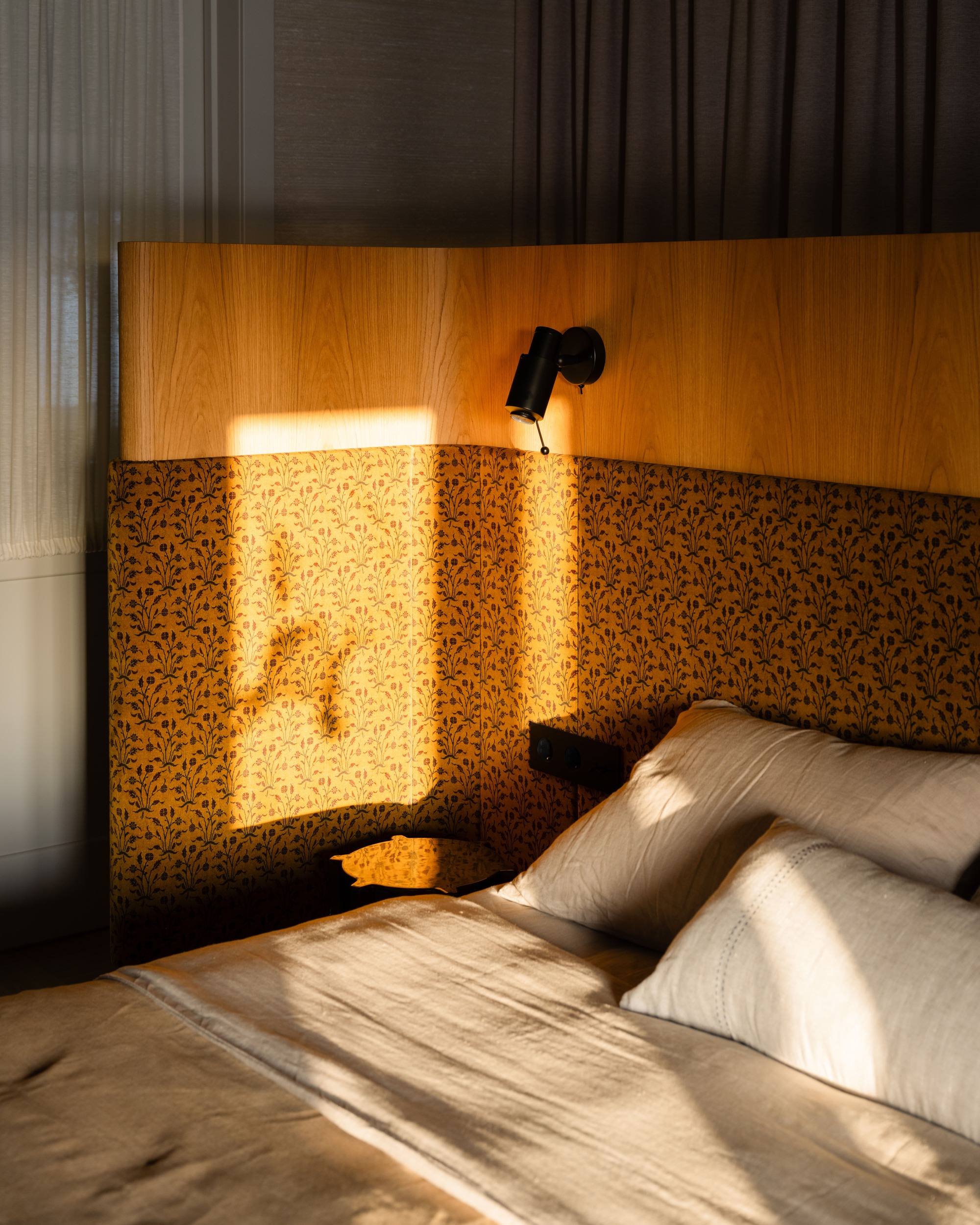 Bed headboard of oak veneer and patterned fabric with a black reading lamp, lit by low sun and a leaf shadow