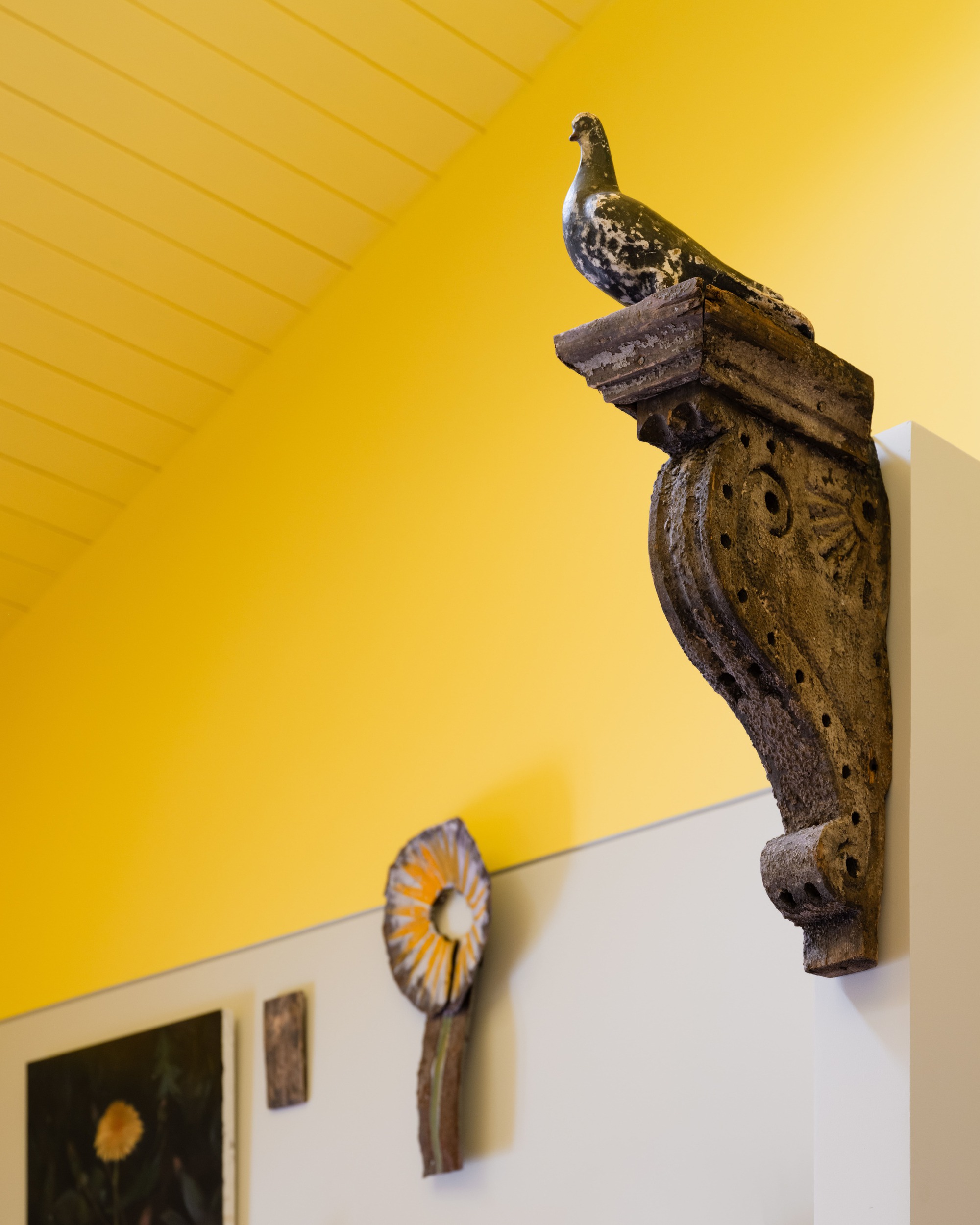 Weathered carved corbel topped with a bird figure against a yellow wall and sloped timber ceiling, with art below