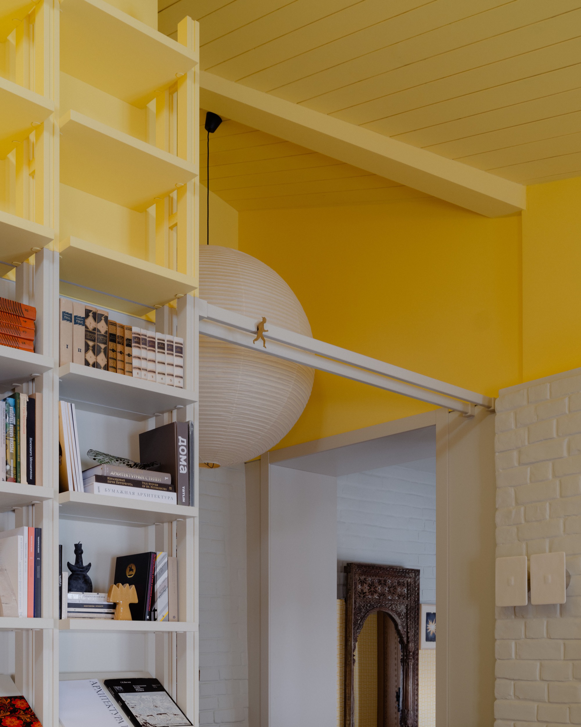 White shelving with books beside a paper globe pendant, a sliding rail and a painted brick doorway under a yellow ceiling