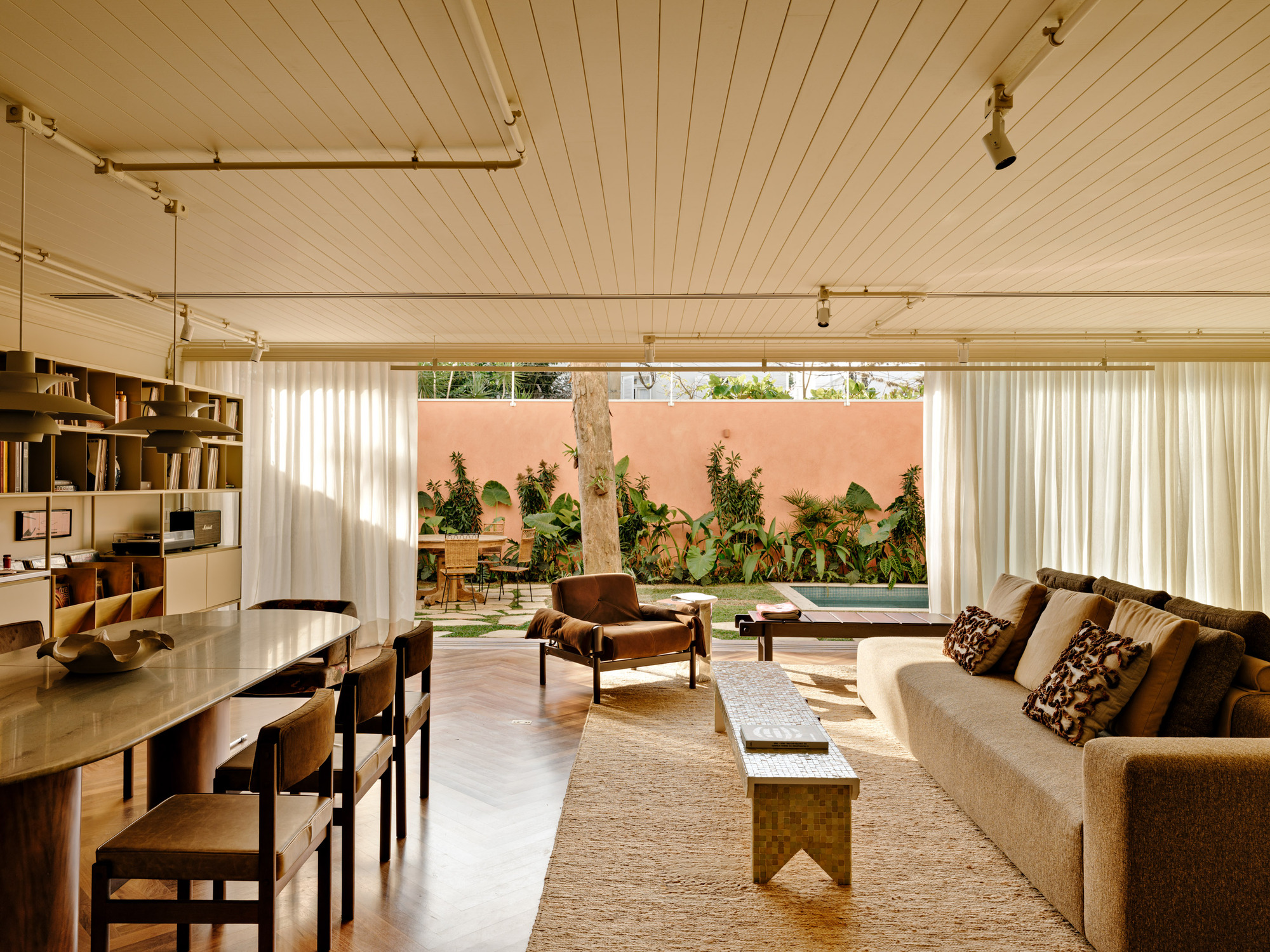 Living and dining room opening through sheer curtains to a pink garden wall, planting and a small pool in afternoon light