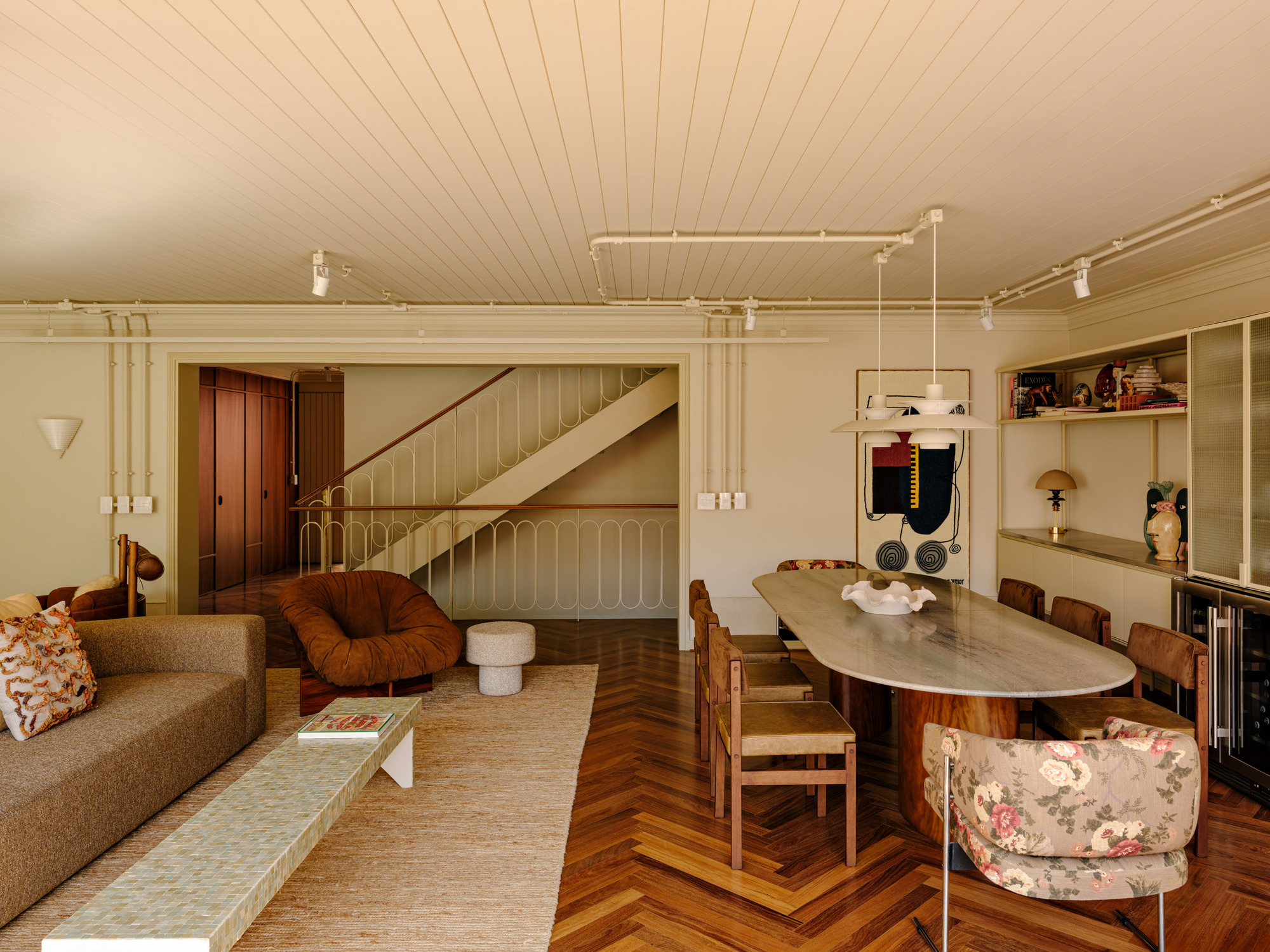 Open living and dining area with a white balustraded staircase beyond, herringbone floor and an oval stone table in warm sun