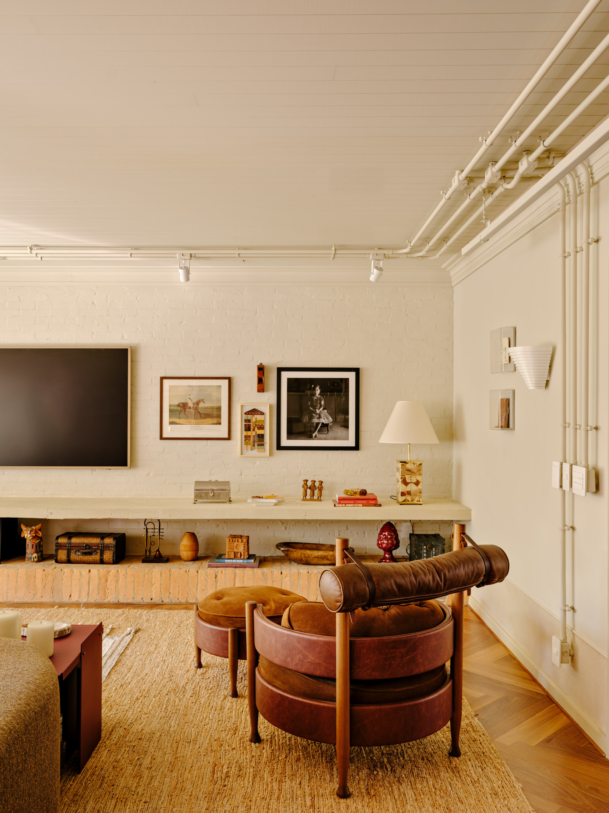 Leather and timber armchair in front of a painted brick wall with framed pictures, a low shelf and exposed white conduits