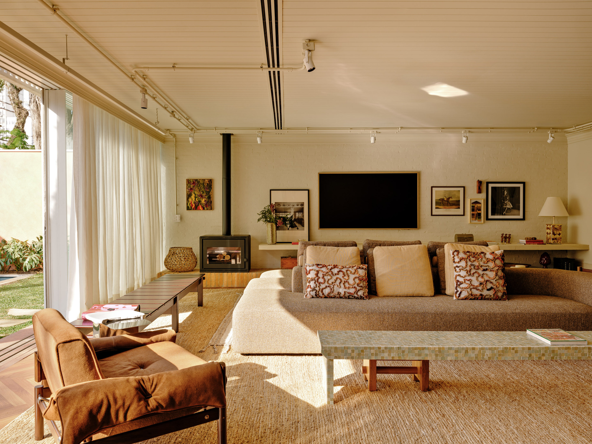 Sitting area with a freestanding black flue fireplace, wall-mounted television and a mosaic-topped bench, beside open sliding glass and sheer curtains