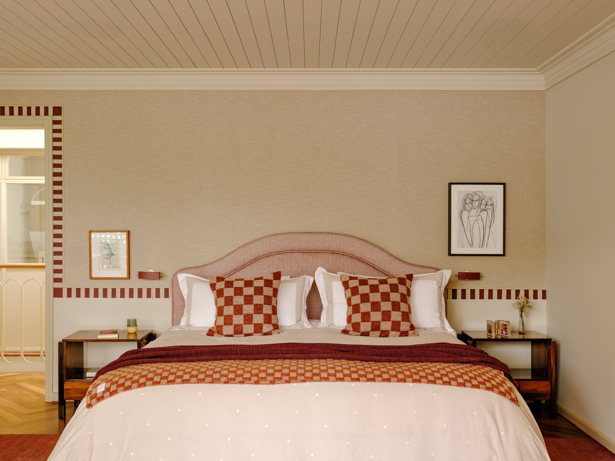 Bed with arched pink headboard and checked cushions against a linen wall with a red dashed border, under a timber-board ceiling