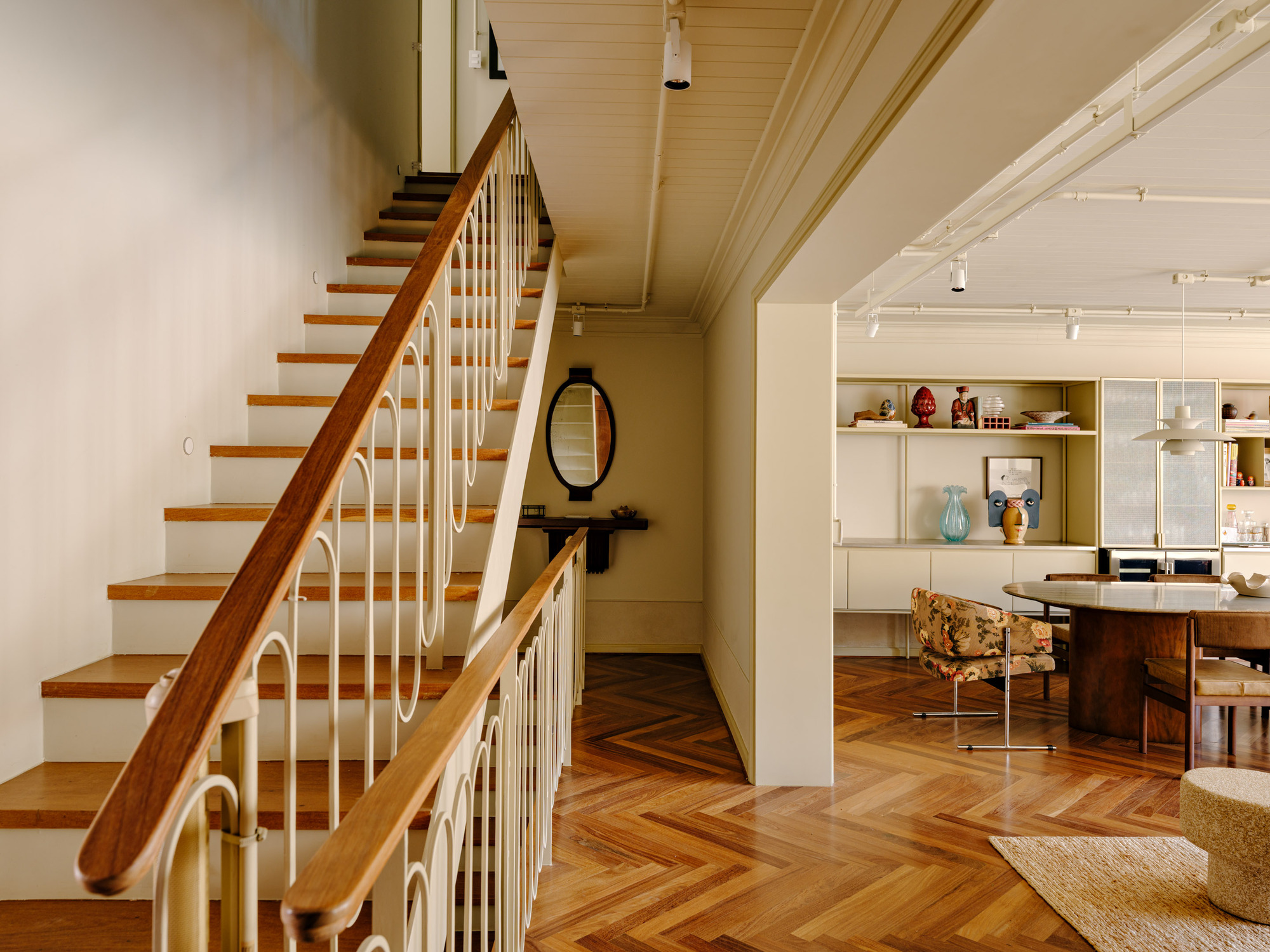 Staircase with timber handrail and looped metal balusters beside a herringbone floor, opening toward a dining area with a round table
