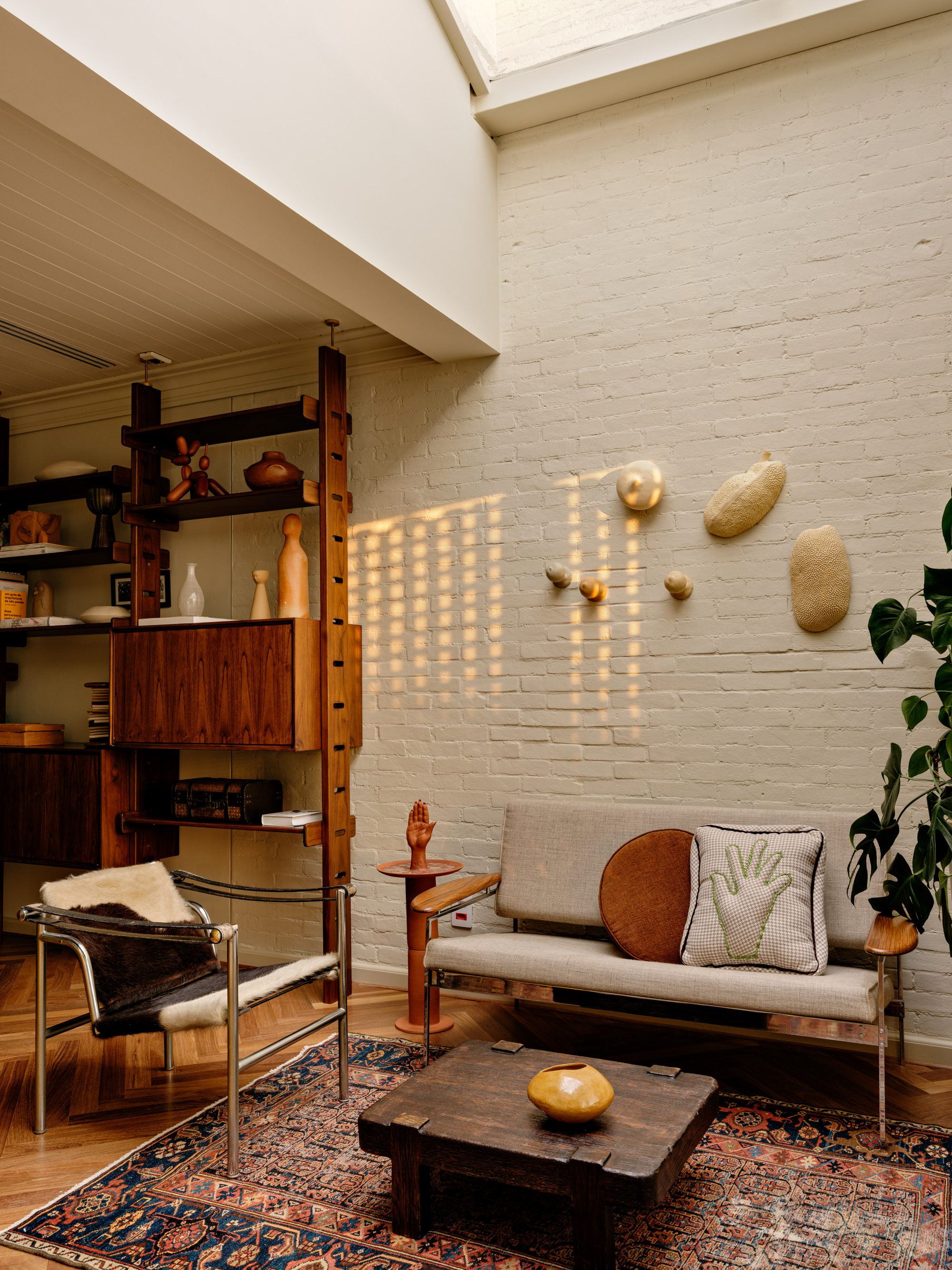 Painted white brick wall with sunlight patches falling across a grey bench, wooden shelving unit and low table on a patterned rug