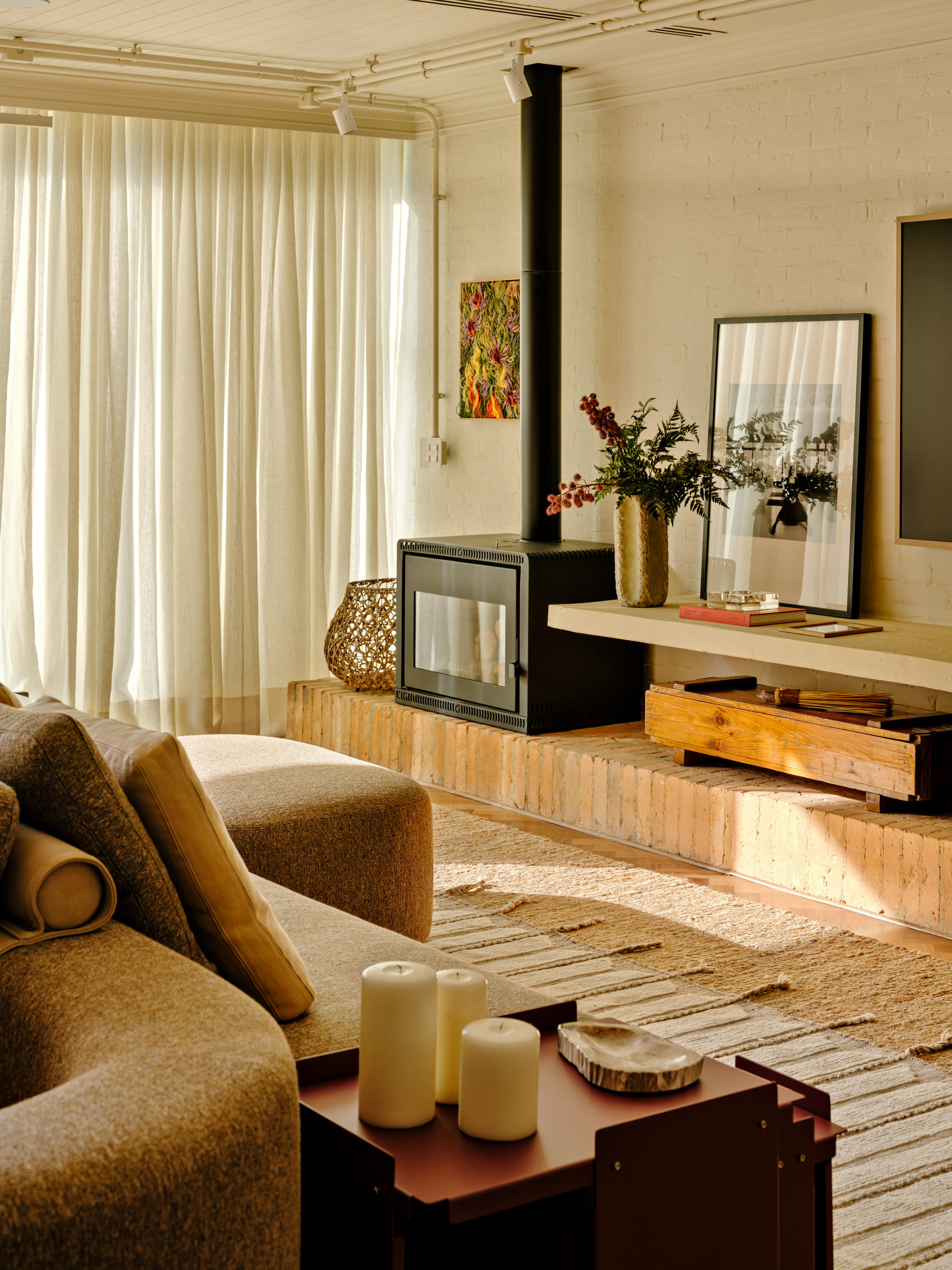 Black wood stove on a raised timber-block platform beside sheer curtains and a curved sofa in warm late light