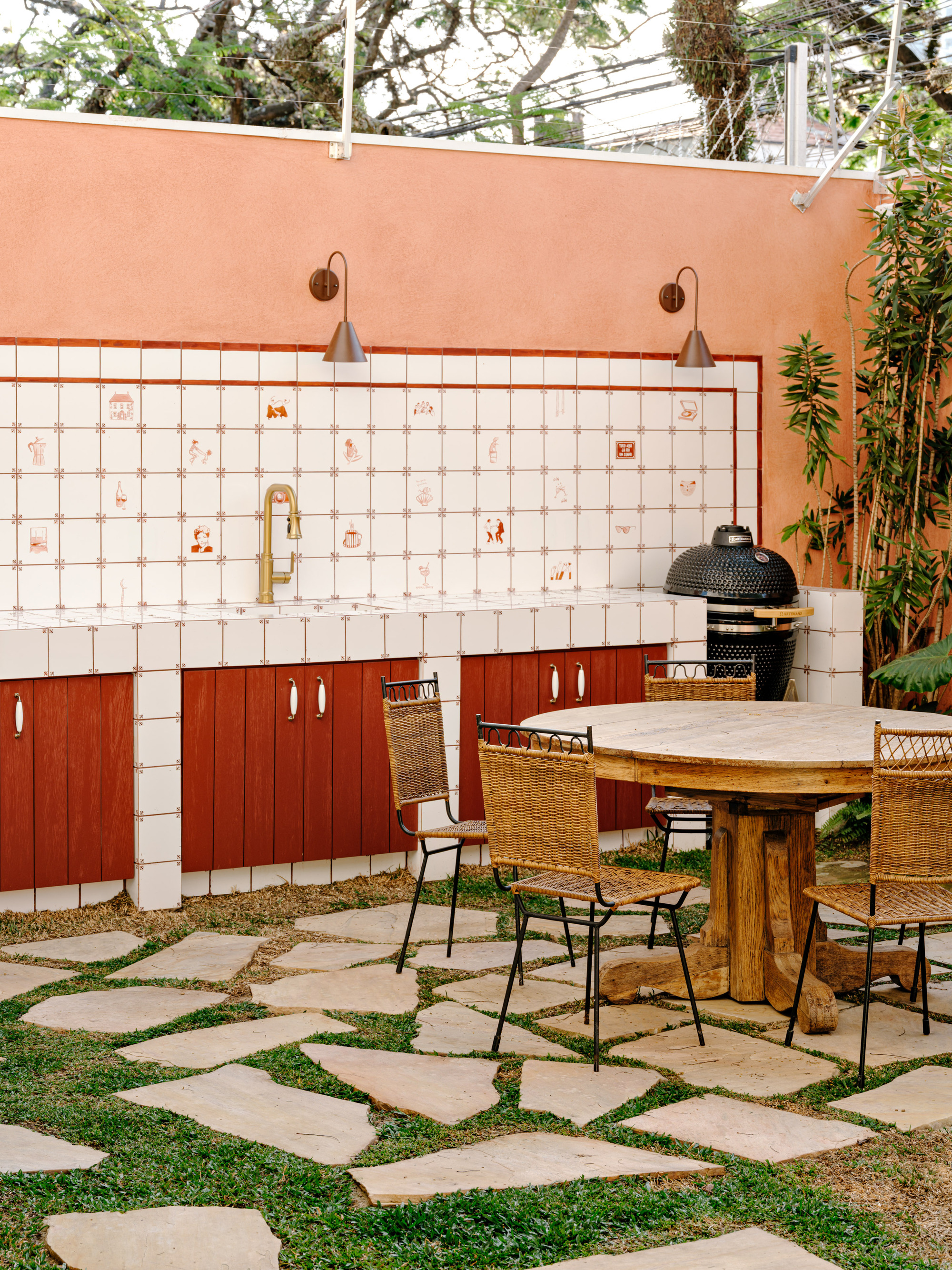 Outdoor kitchen with illustrated white tile backsplash, red cabinet doors and a round wooden table on irregular stepping stones in grass
