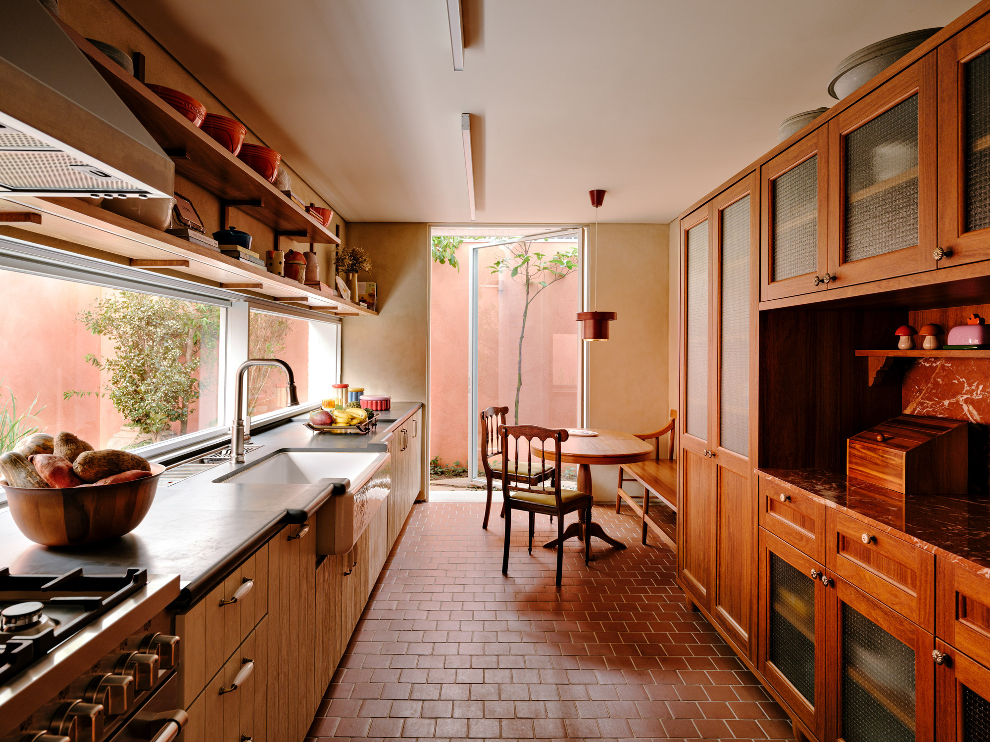 Galley kitchen with timber cabinetry, brick floor and a small dining table facing a pink courtyard wall with a slender tree