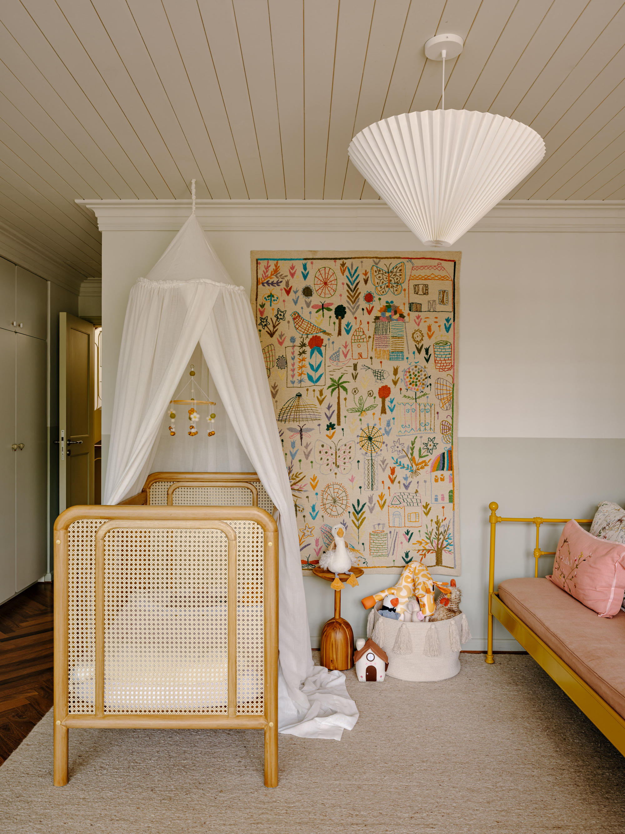 Nursery corner with a rattan crib under a white canopy, an embroidered wall hanging and a pleated cone pendant lamp