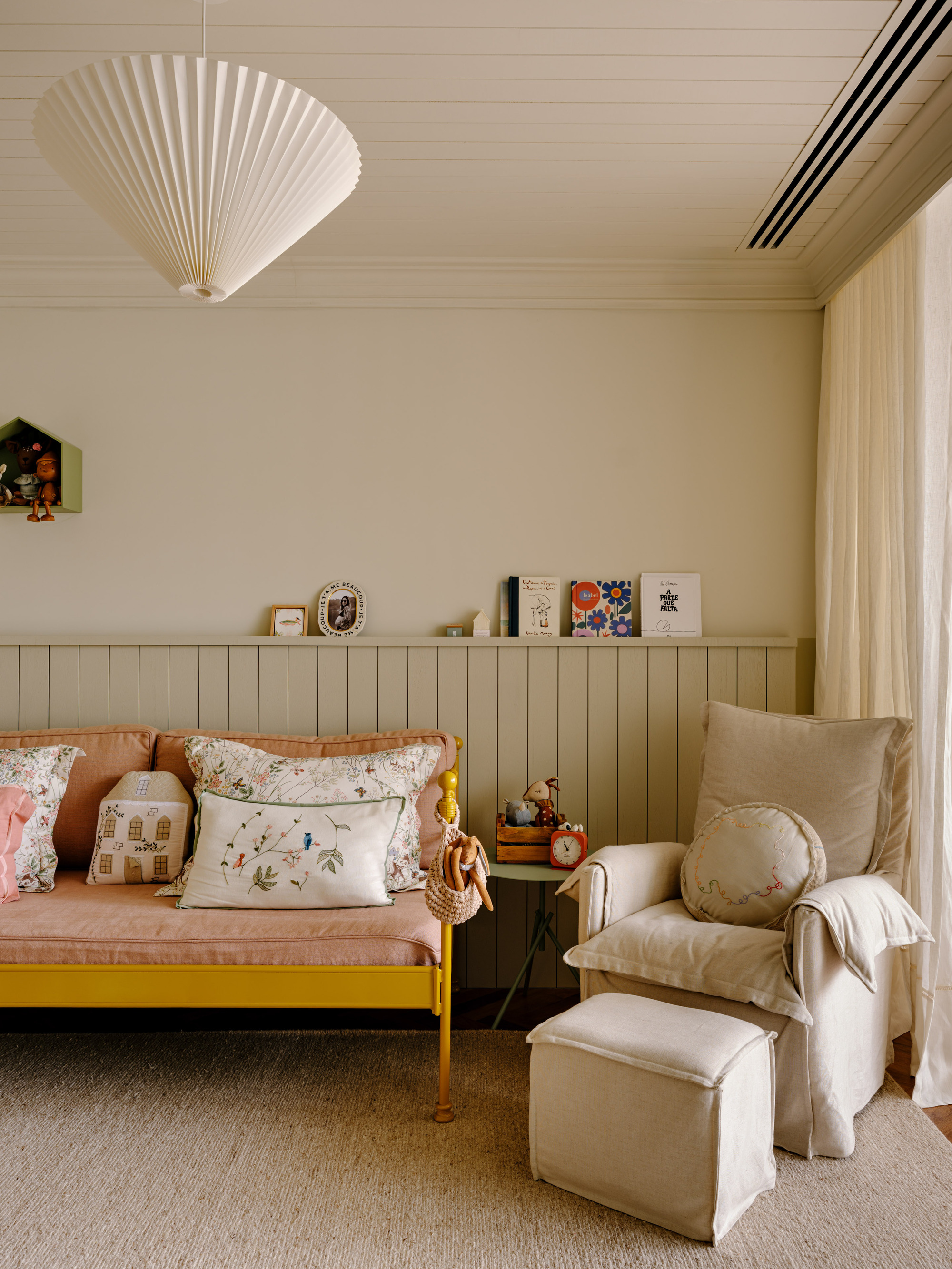 Yellow-framed daybed with floral cushions beside a linen armchair and ottoman, lit by soft daylight through a sheer curtain