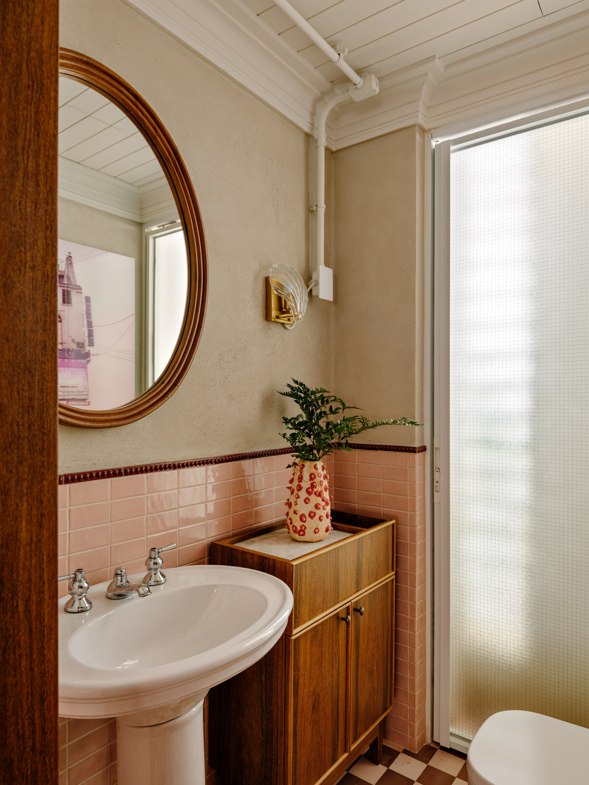 Powder room with a pedestal sink, pink tile wainscot, oval wood-framed mirror and a spotted vase holding a fern sprig