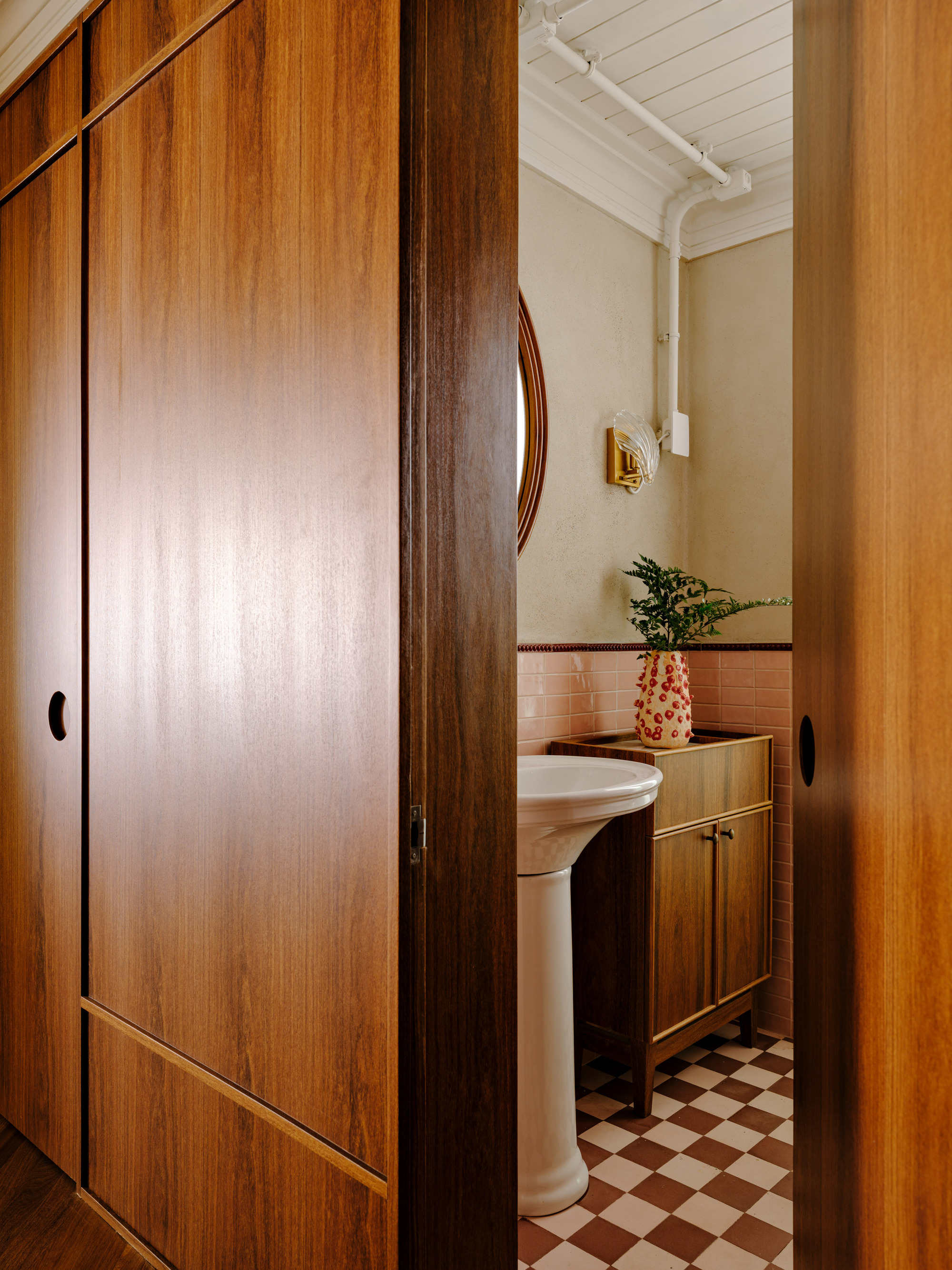 View through sliding timber doors into a small bathroom with a checkered floor and pedestal sink, seen in warm light