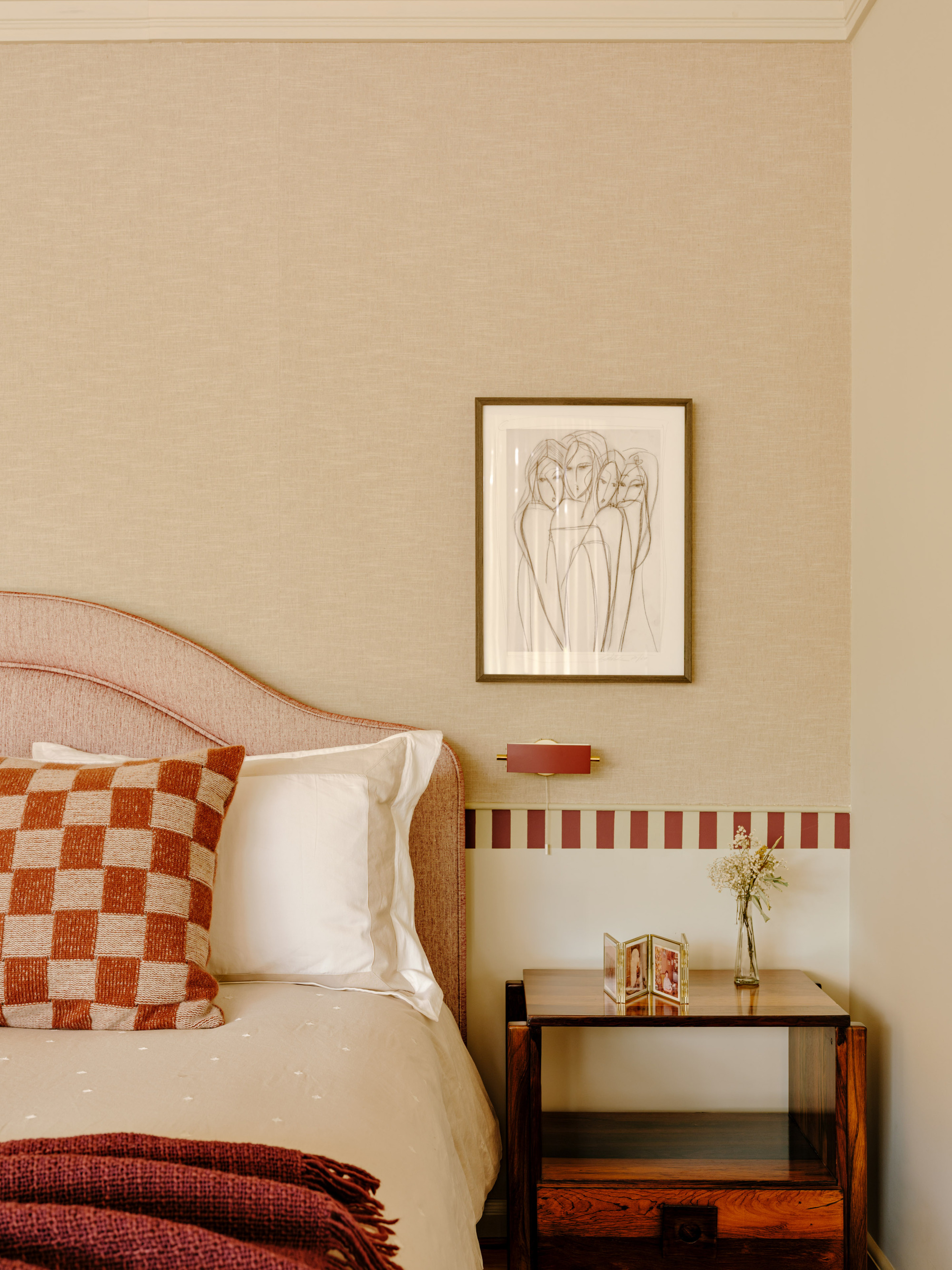 Bedside corner with an arched upholstered headboard, checkered cushion, framed line drawing and a red-striped tile border in soft daylight