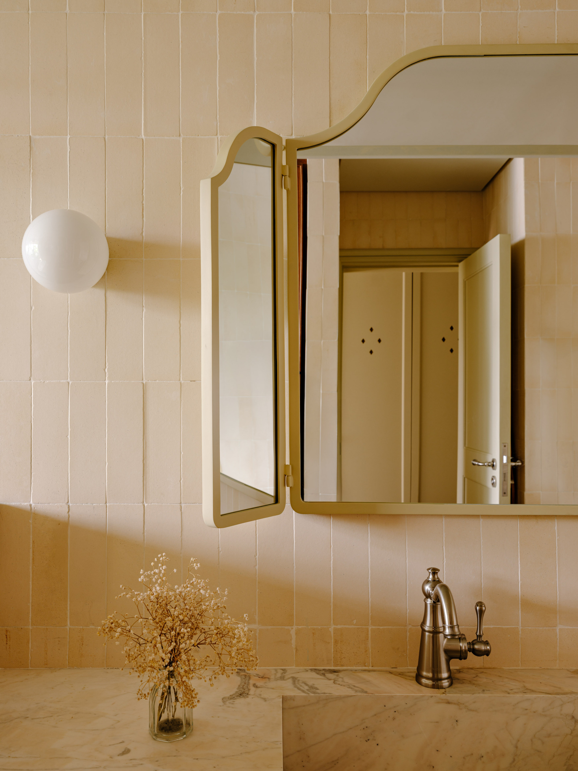 Folding arched mirror cabinet on tiled wall beside a globe sconce, with dried flowers in a glass vase on the marble basin