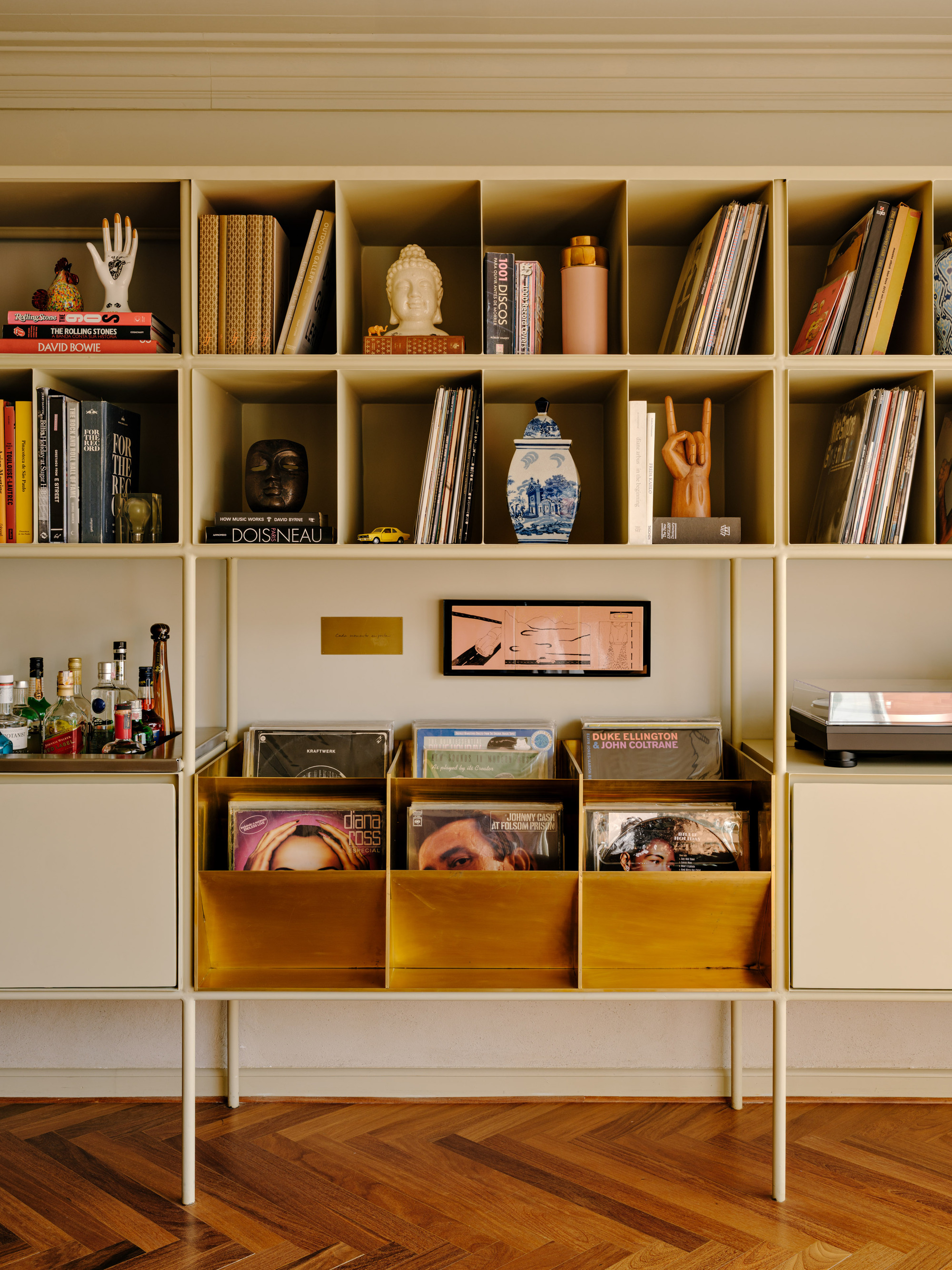 Cream metal shelving unit holding vinyl records in brass bins, a drinks tray and ceramics above a herringbone timber floor