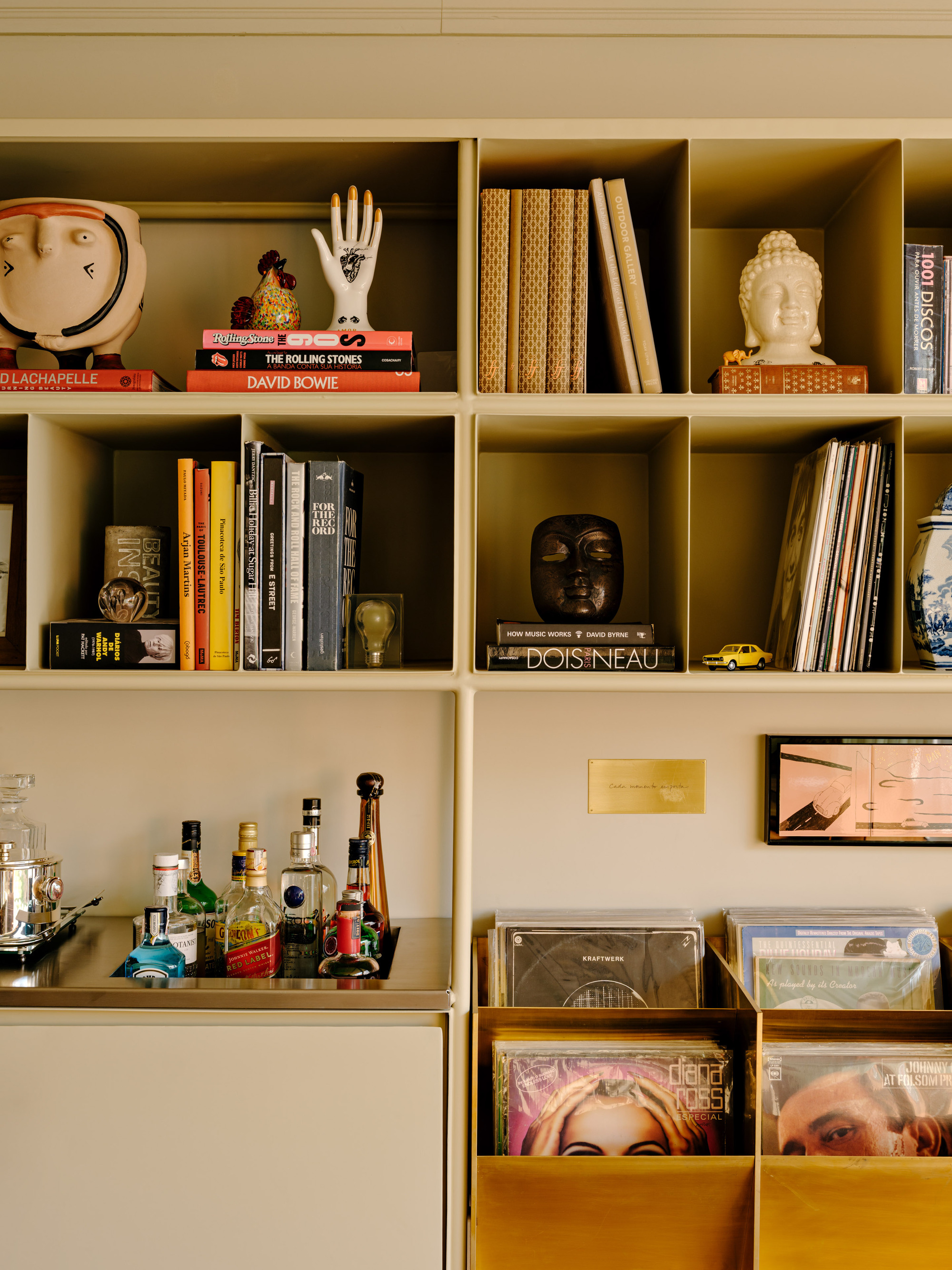 Close-up of cubby shelves with stacked books, a white hand sculpture, a Buddha head and bottles on a steel bar tray