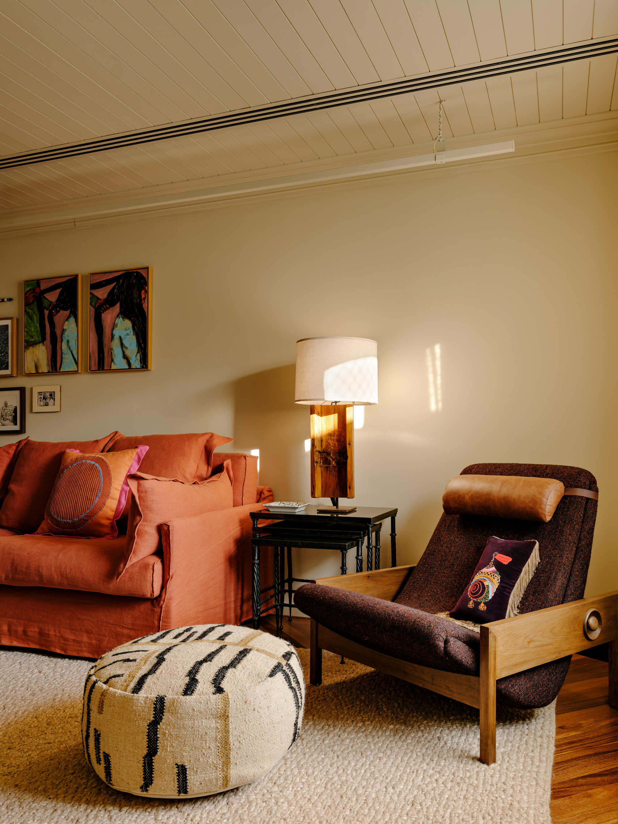 Timber lounge chair, round woven pouf and wood-based table lamp beside the sofa, lit by low raking sun