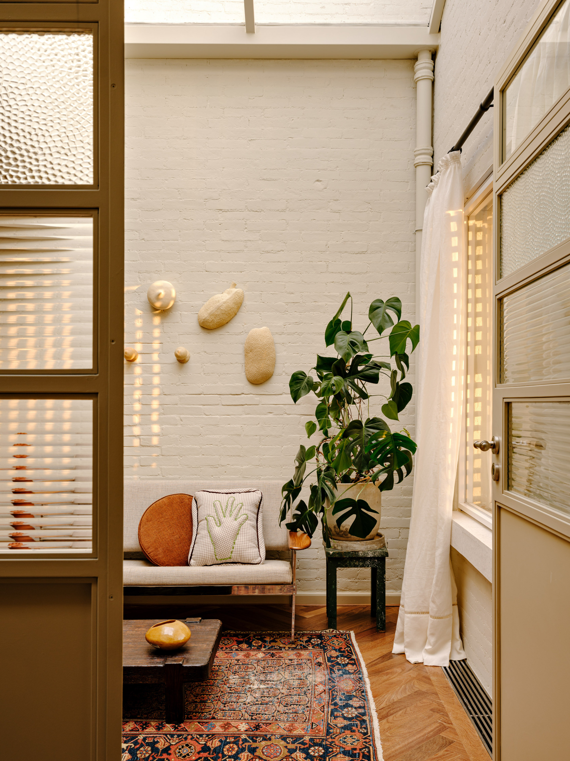 Painted white brick wall with ceramic wall pieces, a monstera plant and a daybed behind reeded glass doors