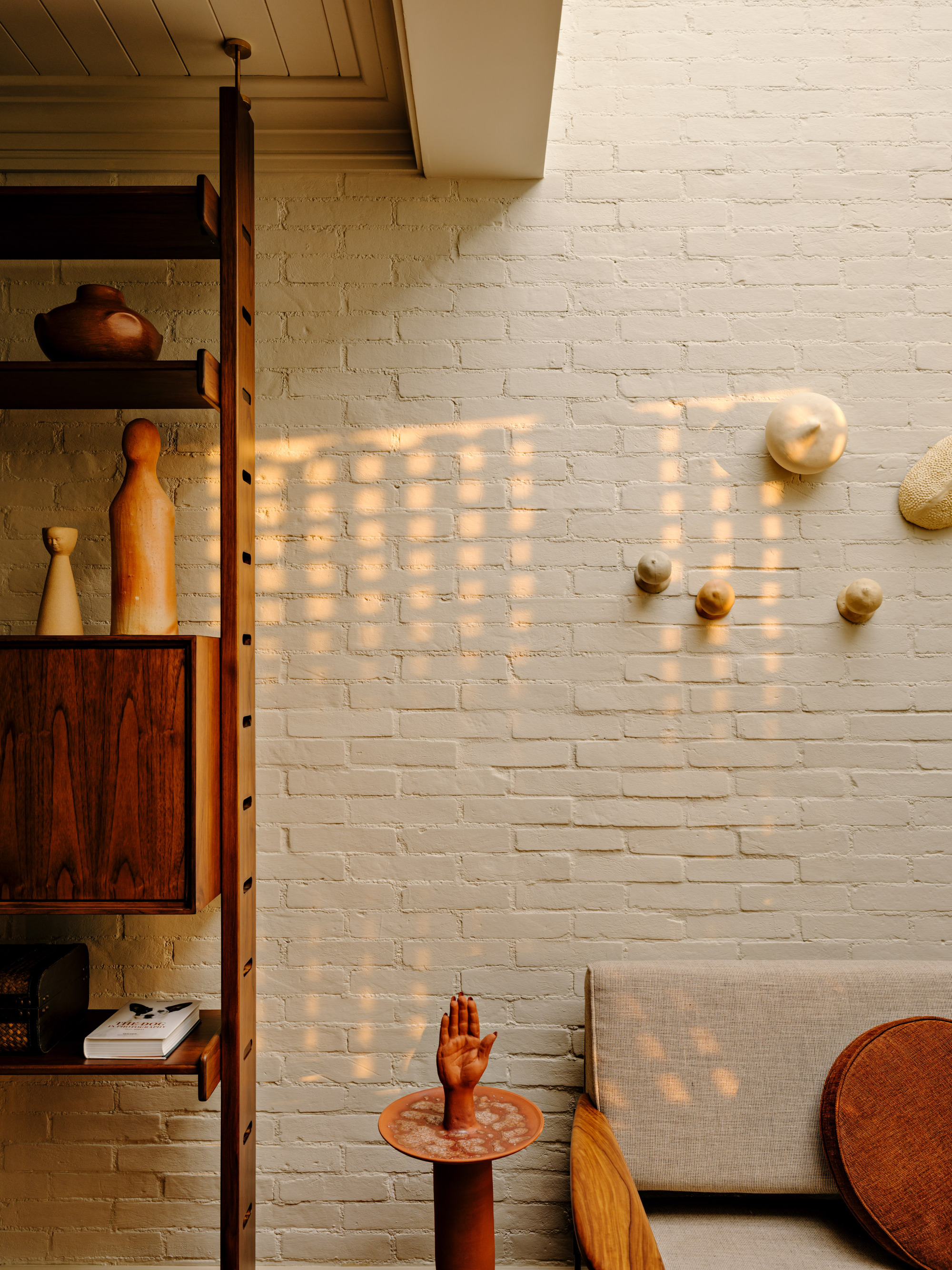 Dappled window light across painted brick, beside a teak shelving unit and a terracotta hand sculpture on a side table