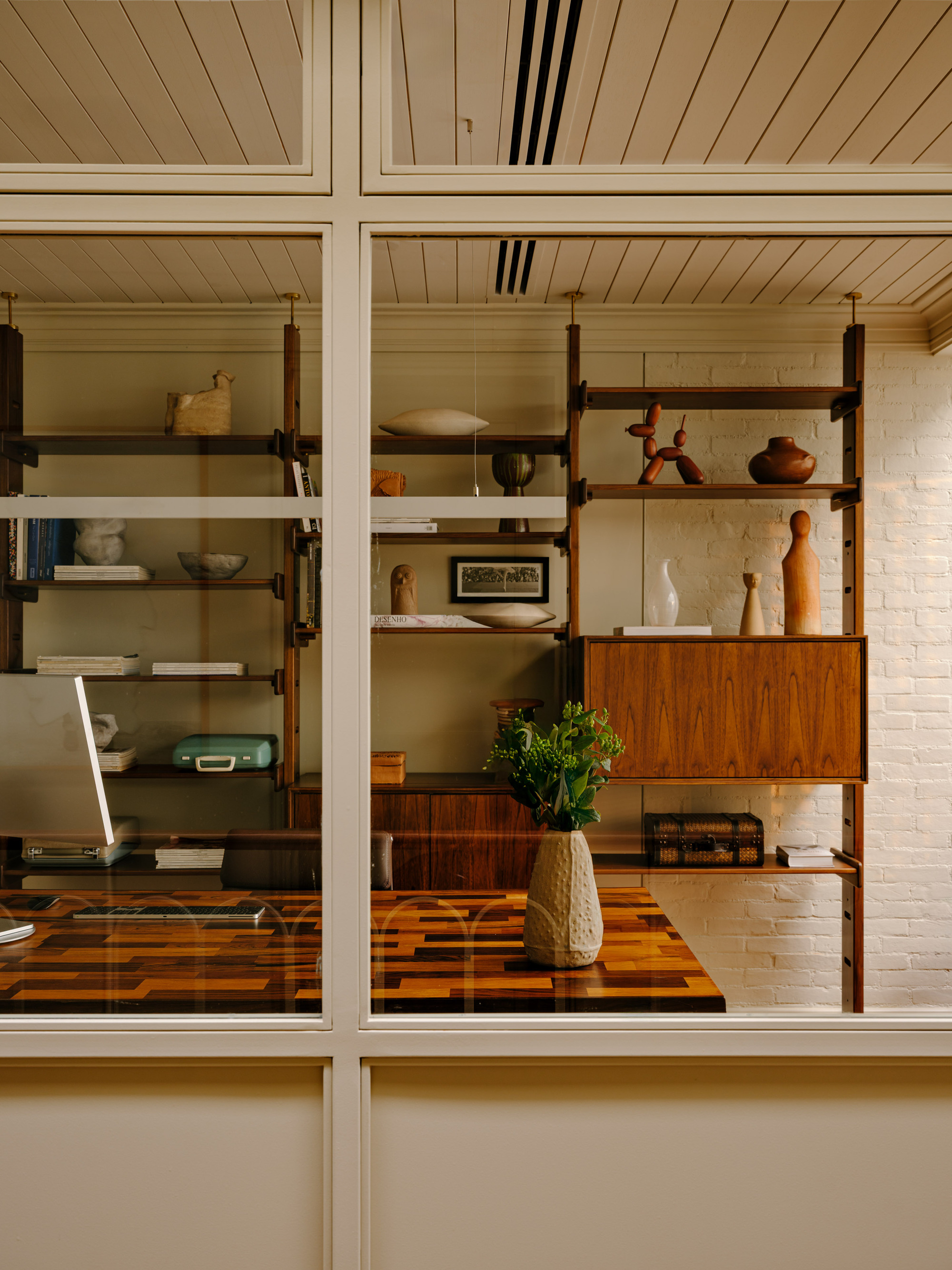 View through a glazed partition to teak shelves and a parquetry desk topped with a vase of greenery