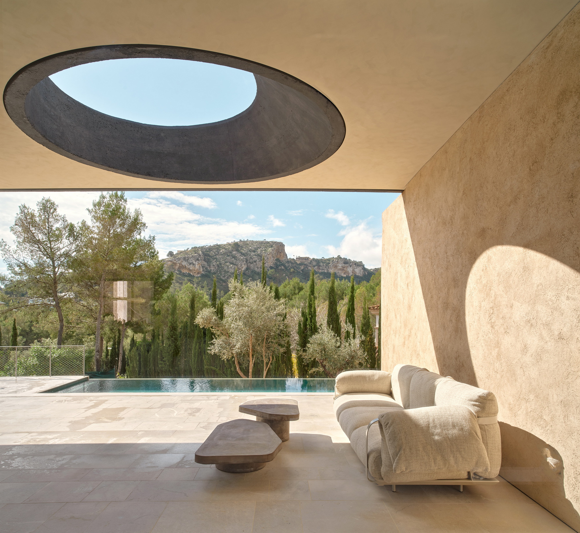 Covered terrace with a round skylight above a cream sofa and low tables, sunlight casting a curved shadow on the plaster wall
