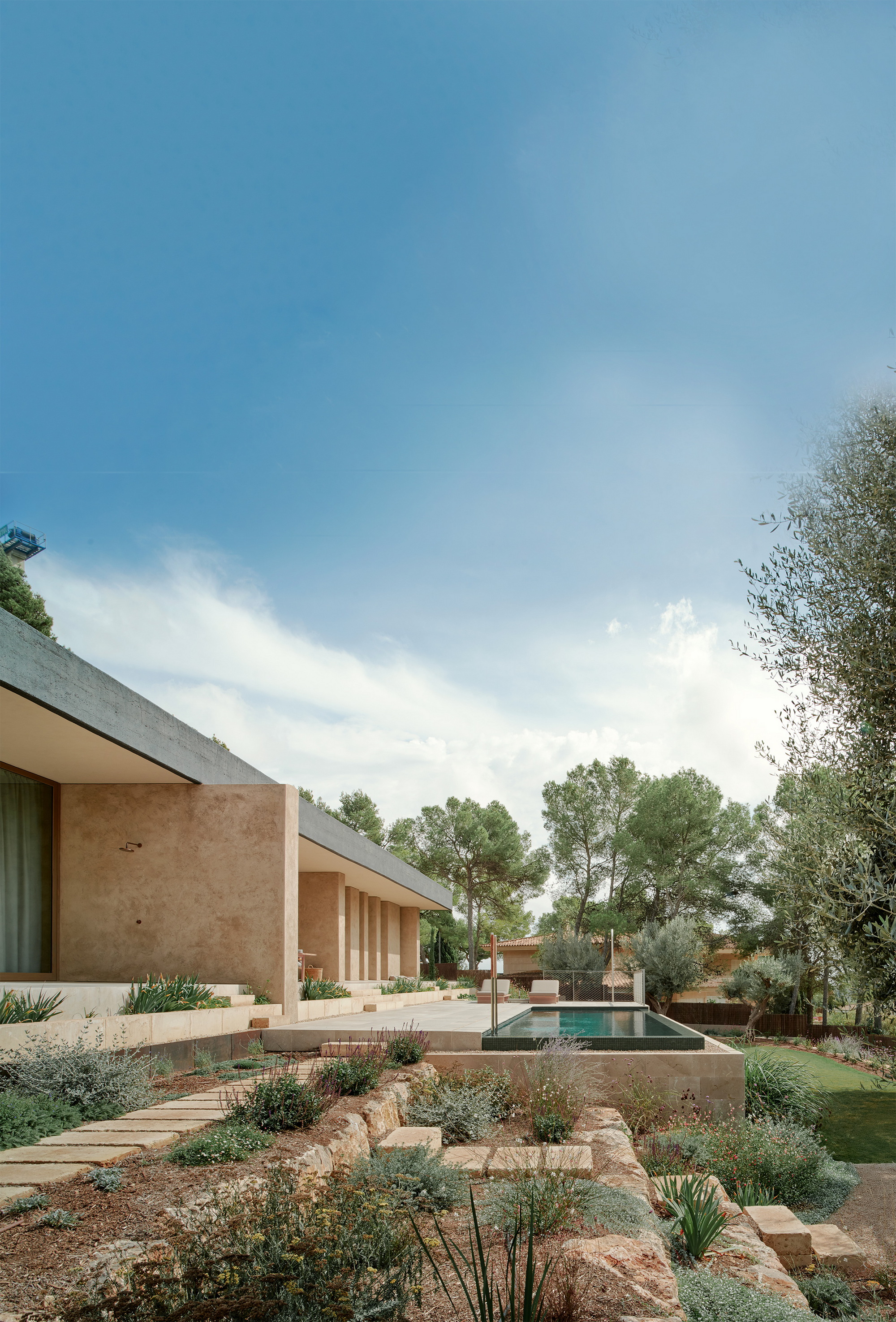 Rammed earth walls and a flat concrete roof edge above a terraced stone garden, with a pool beyond under scattered clouds