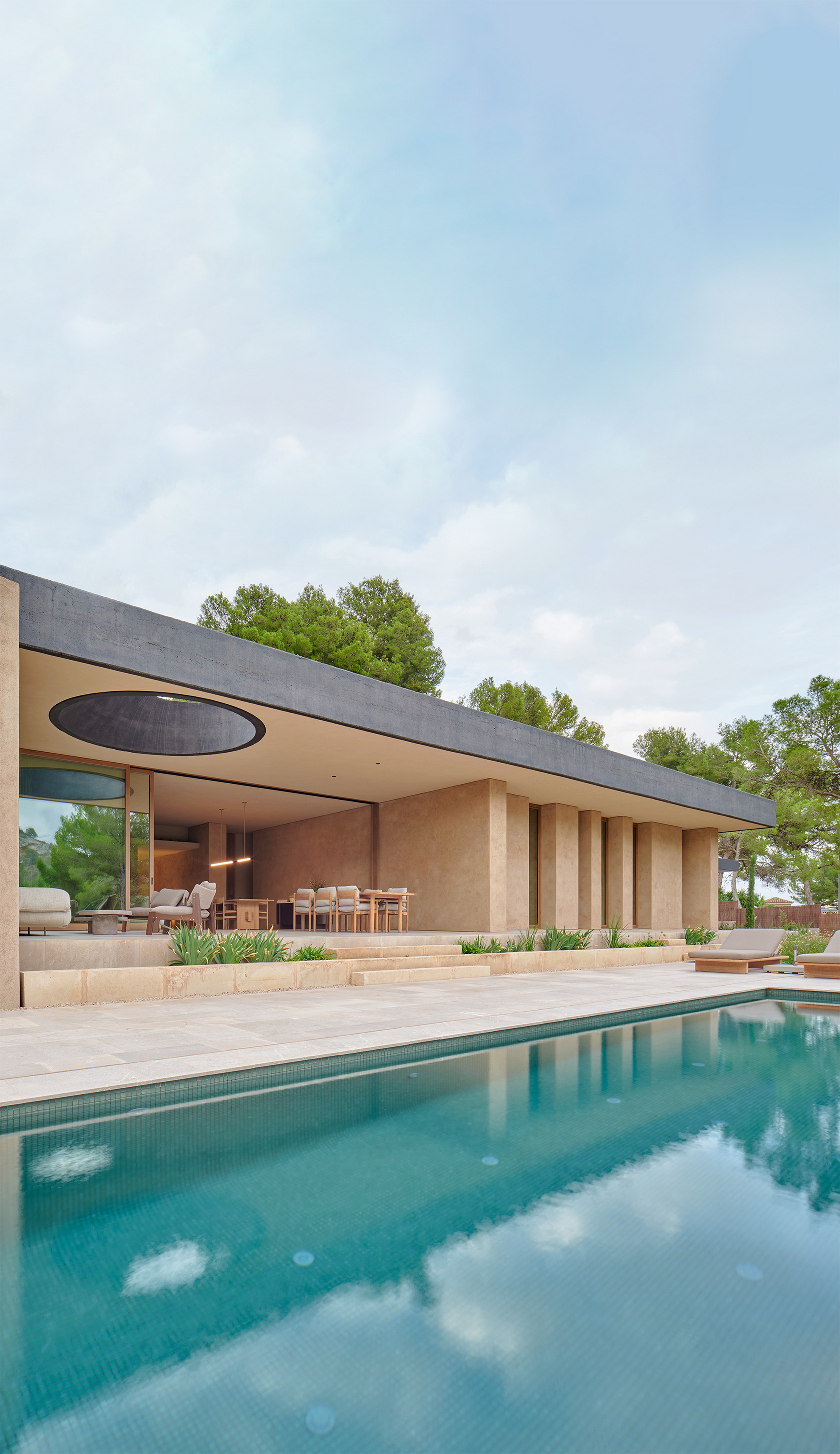 Pool edge in front of a pavilion with earth-toned columns and a round roof opening, pine trees behind in soft cloud light