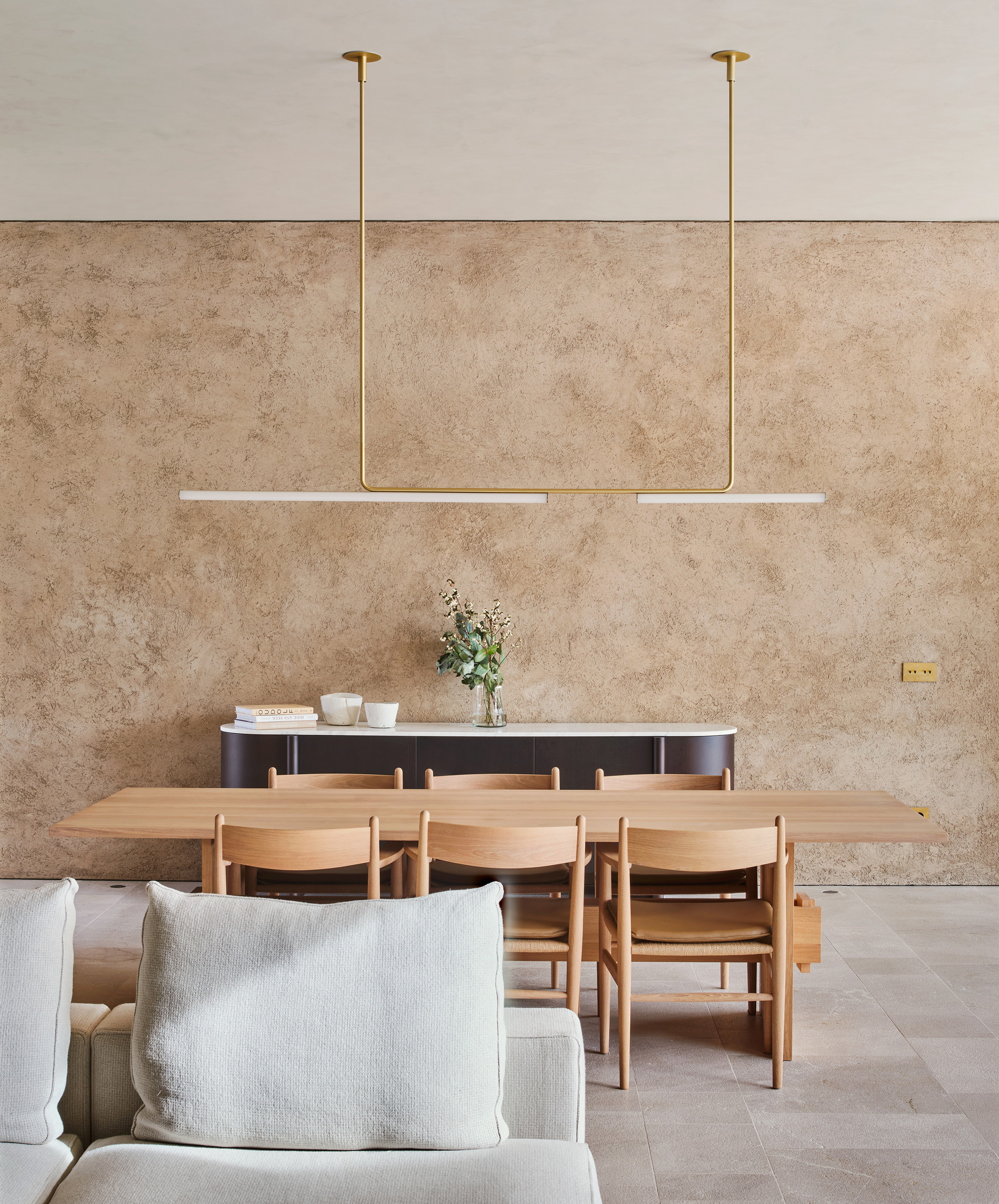 Long timber dining table with oak chairs below a brass linear pendant, set against a textured earthen wall in daylight