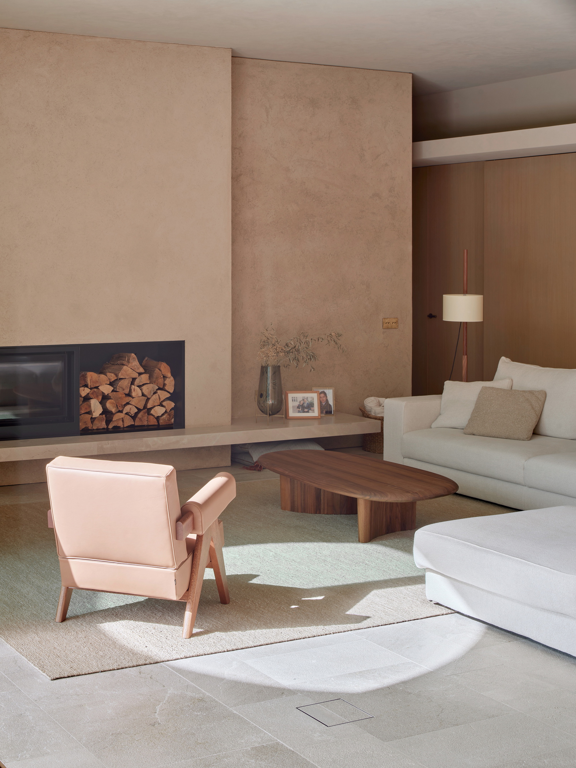 Living room with a recessed fireplace holding stacked logs, a pink leather armchair and a walnut coffee table in a patch of sunlight