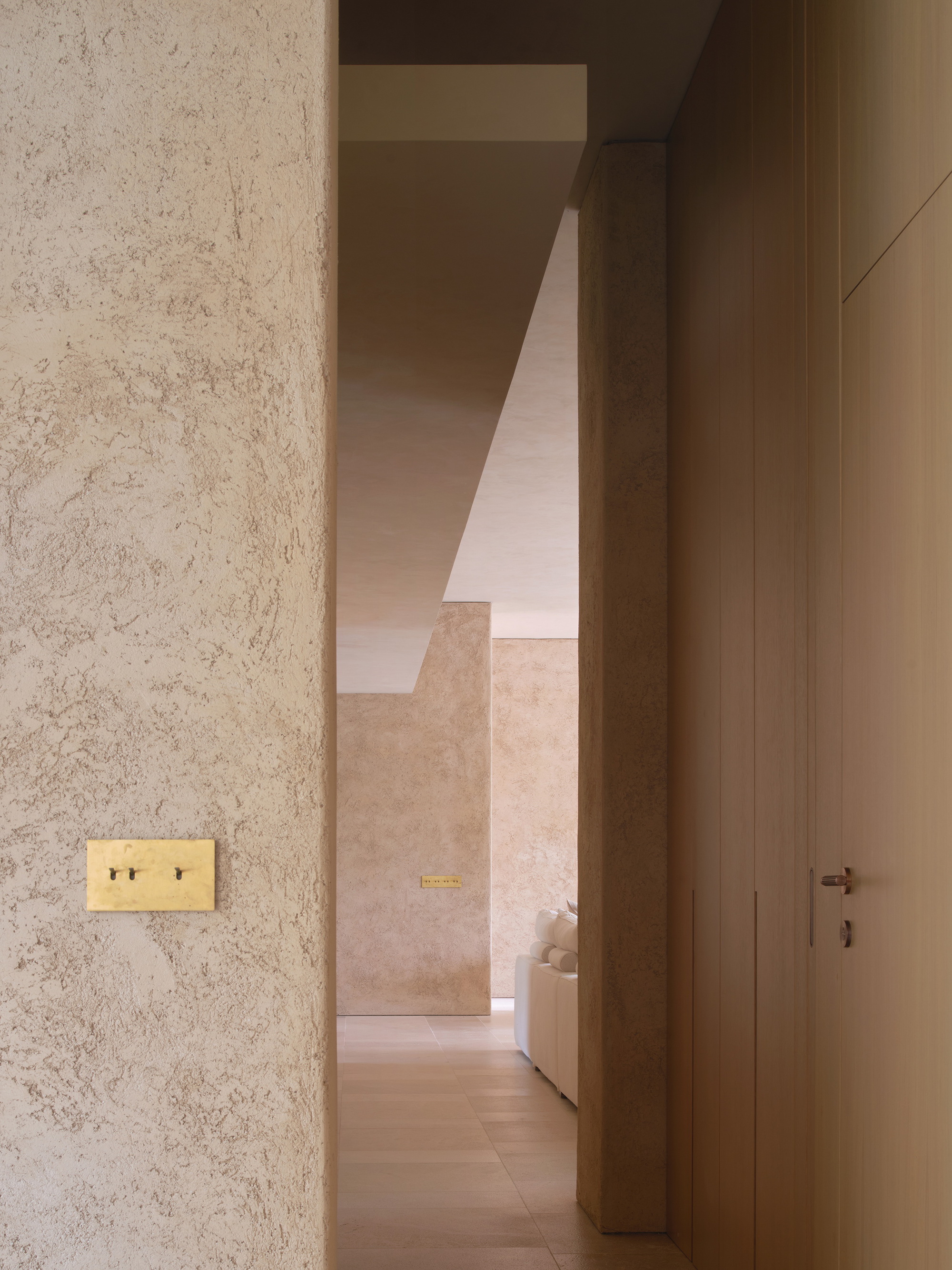 Corridor view past a rough plastered wall with a brass toggle switch plate, toward a sofa in a sunlit room