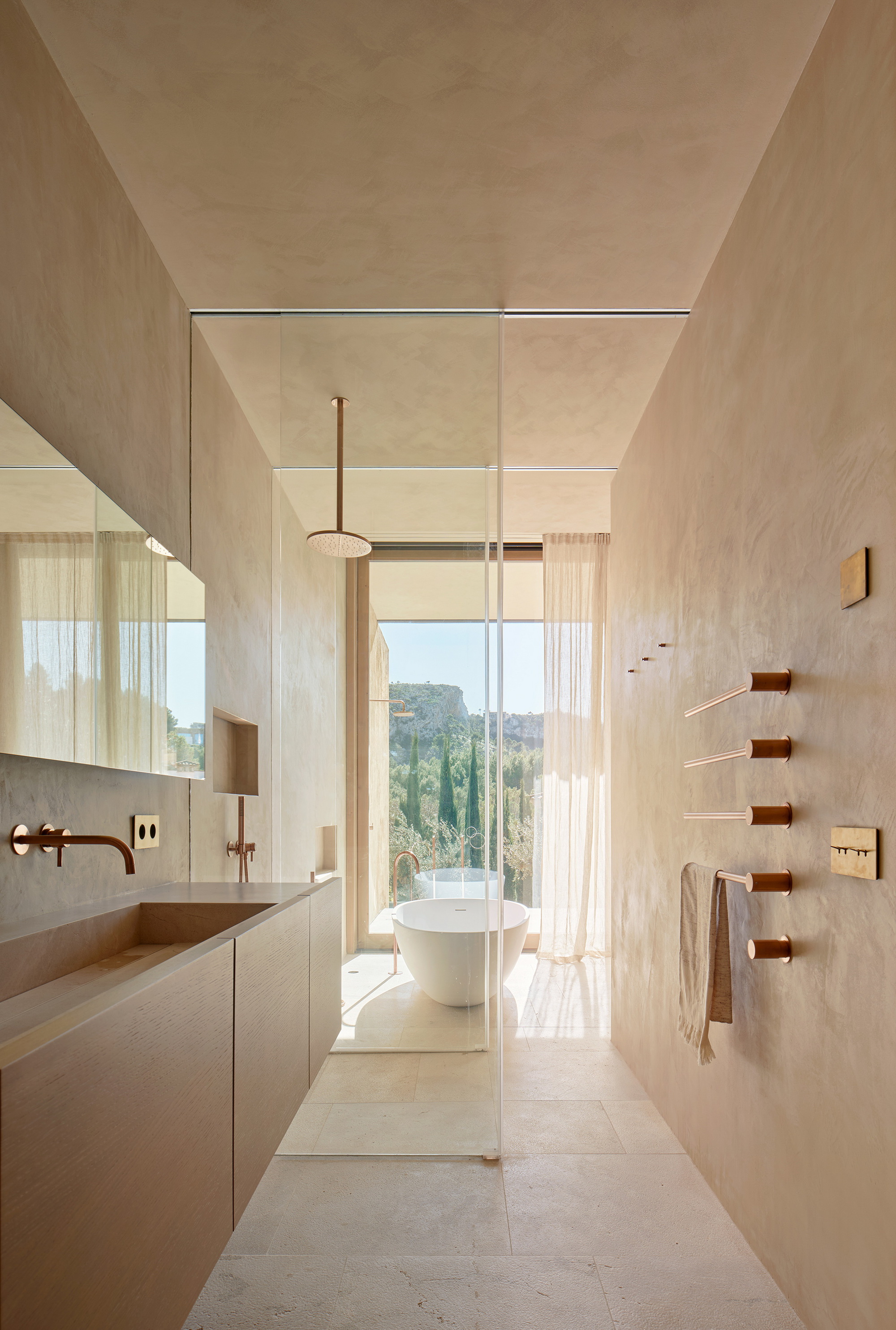 Narrow bathroom with a floating wood vanity and copper fittings, a glass screen opening onto a freestanding tub and cypress trees outside