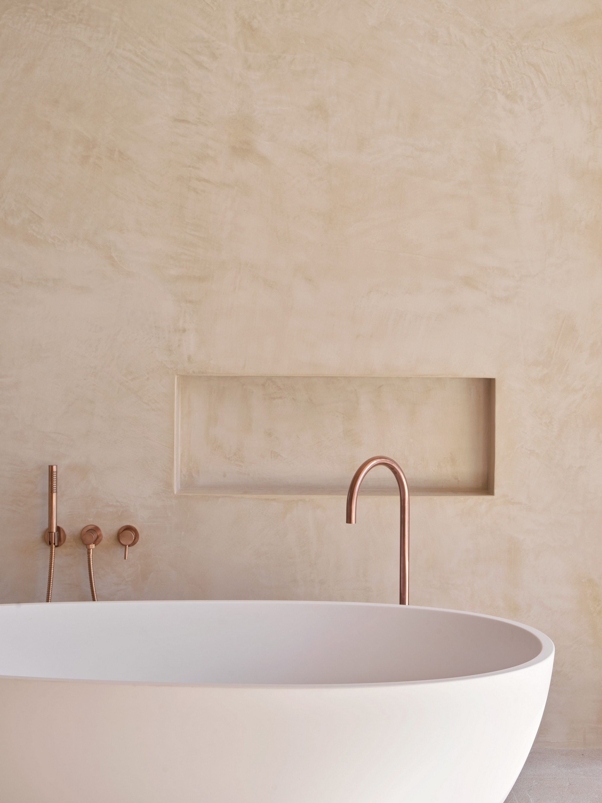Freestanding white oval bathtub with copper tap and wall-mounted controls against a plastered wall with a recessed niche in soft daylight
