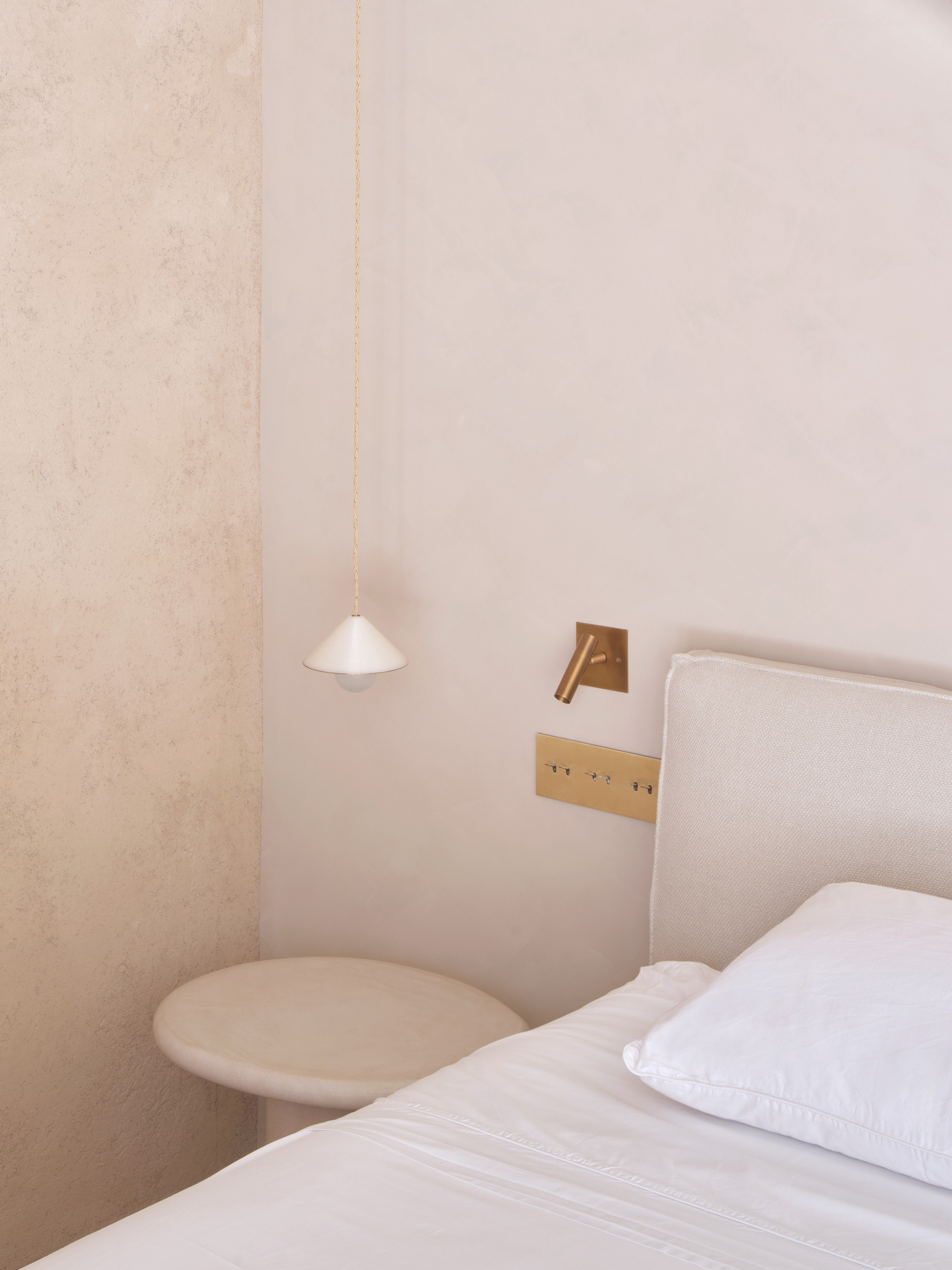 Bedside corner with a cone pendant lamp, brass reading light and round stone side table beside a linen headboard