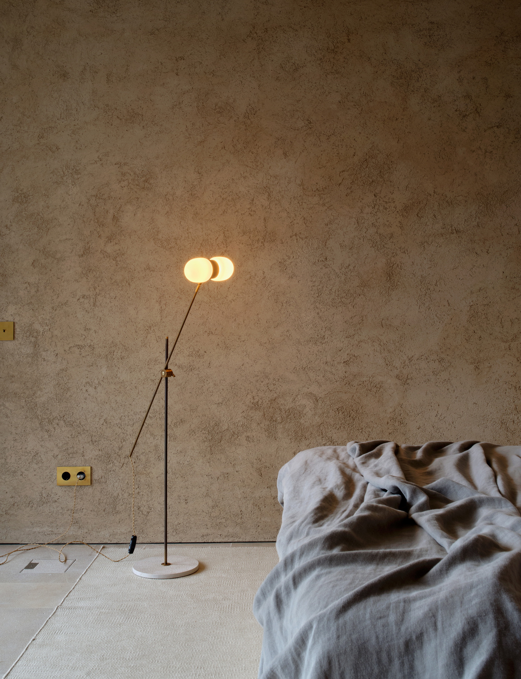 Floor lamp with two glowing globes stands against a rough earthen plaster wall beside a rumpled grey linen bed
