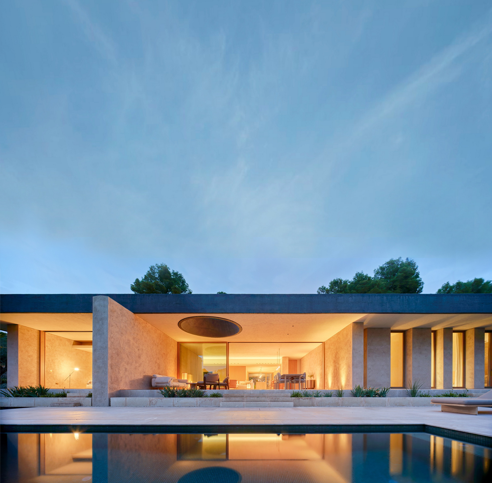 Single-storey pavilion with a flat dark roof and a round skylight, lit from within at dusk beside a still pool