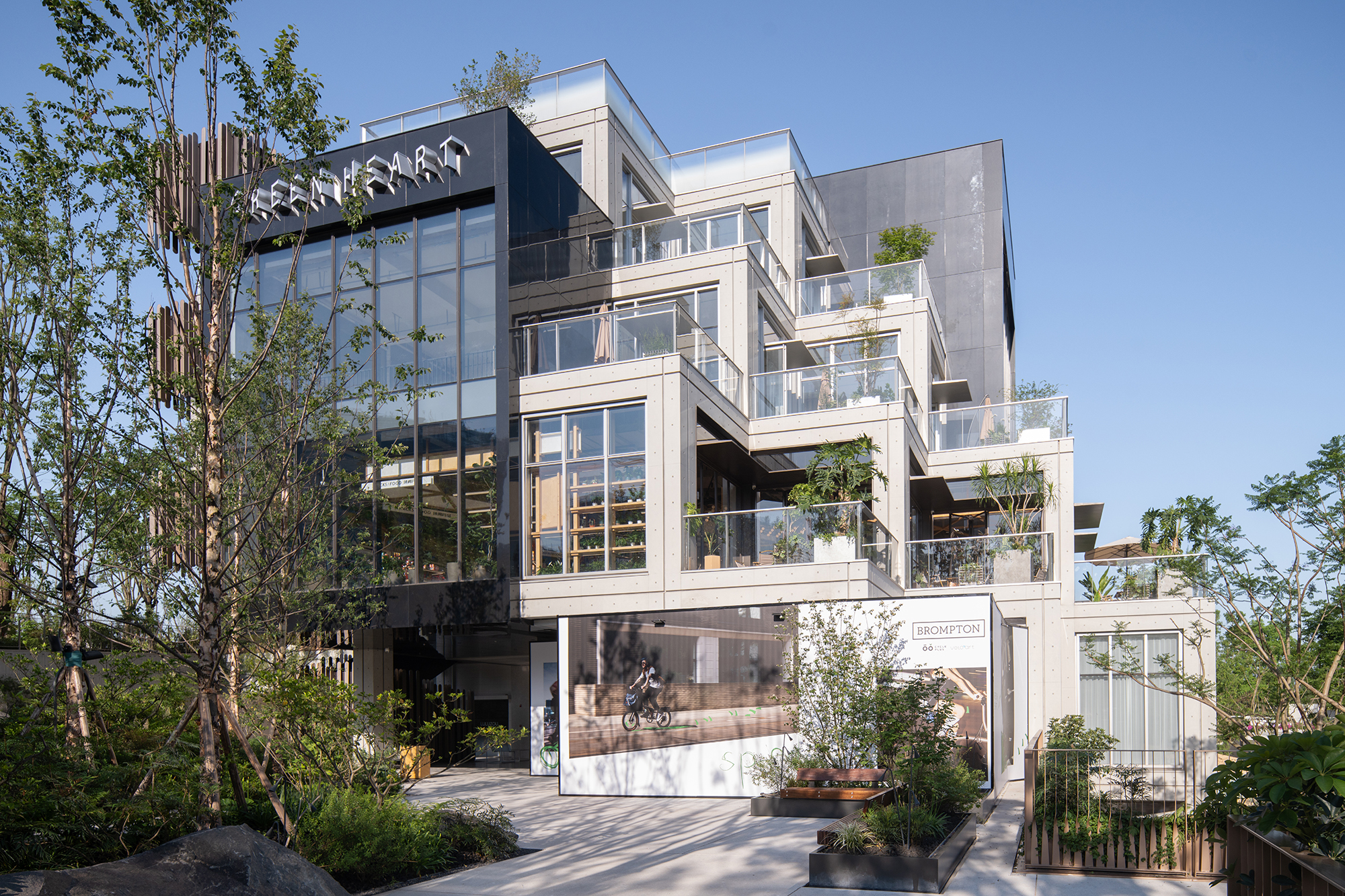Stepped facade of pale stone cubes with glass balustrade terraces and planters, seen through young trees under a clear blue sky