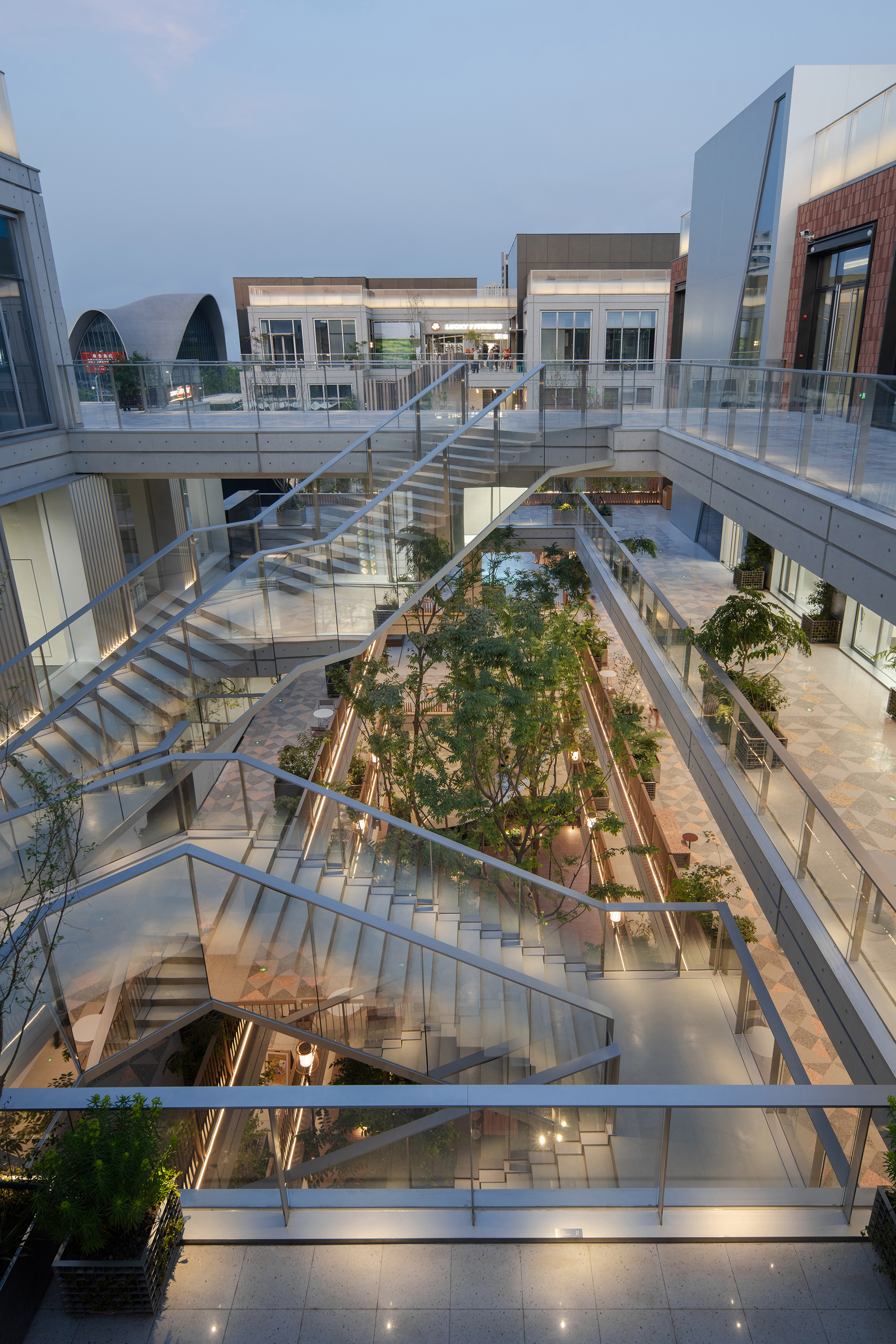 Looking down into an atrium with glass-railed stairs, bridges and a central tree planting, lit by warm lights at dusk