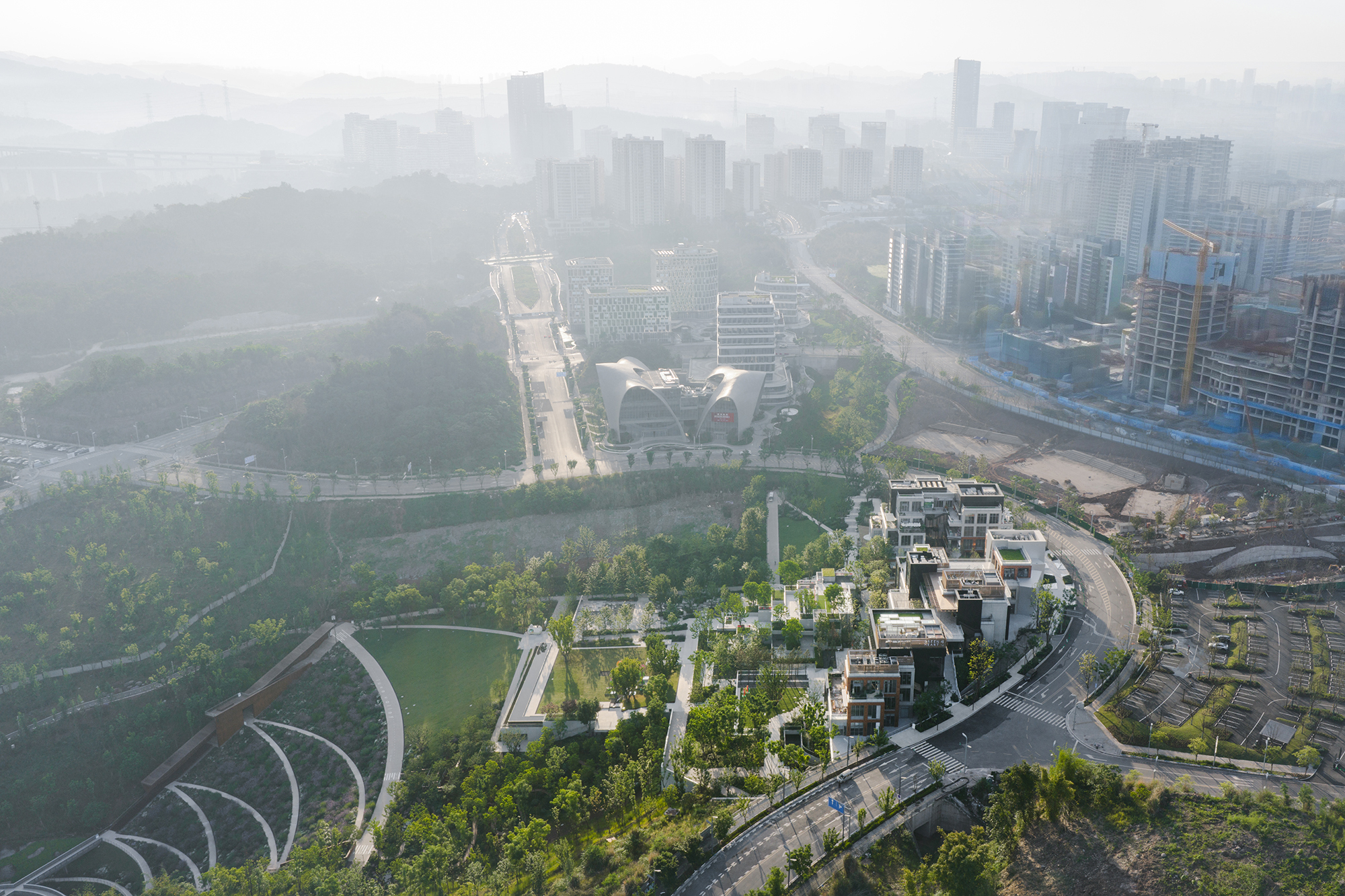 Aerial view of terraced stone-edged gardens and stepped blocks beside a curving road, with towers rising through morning haze