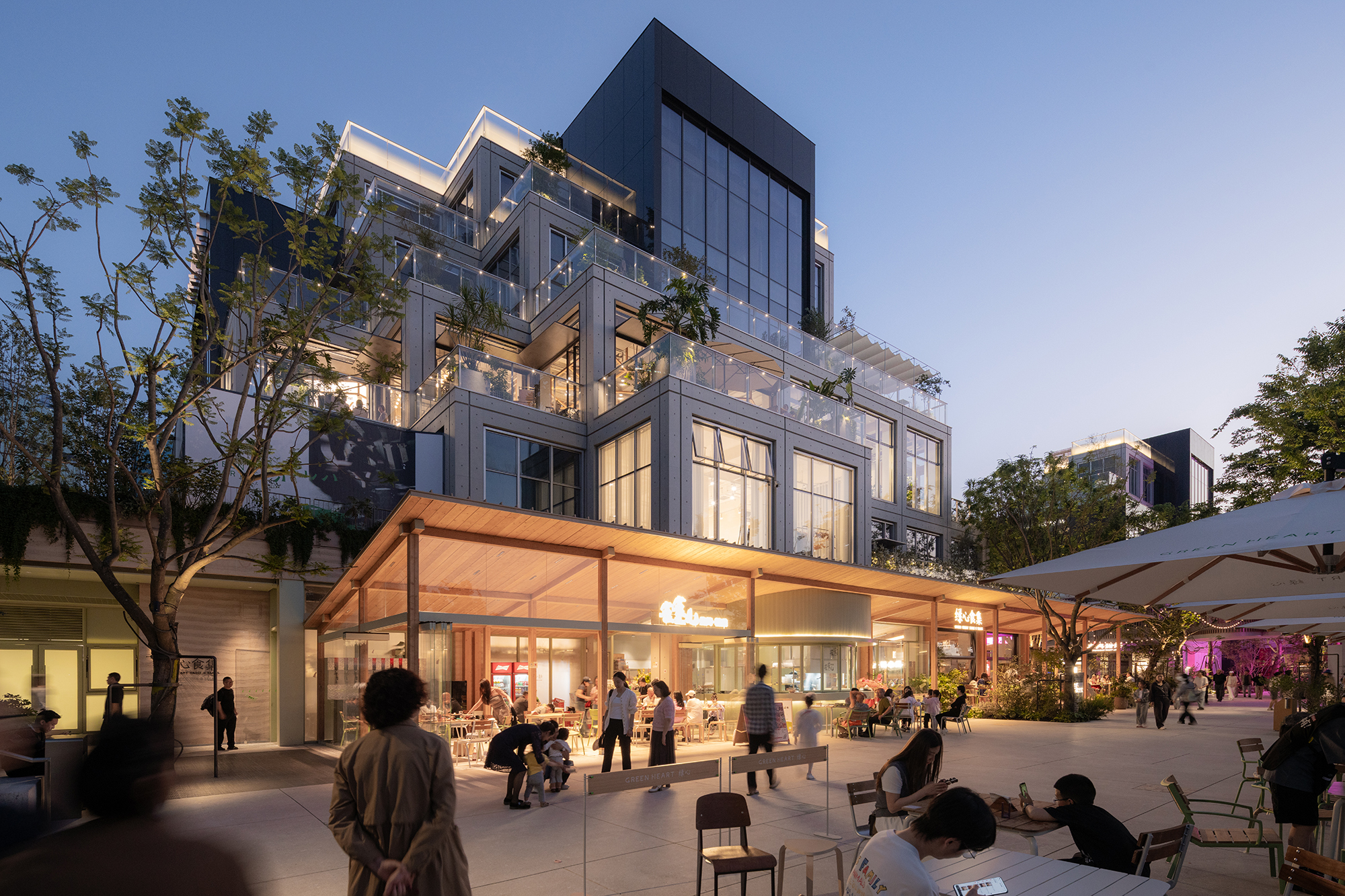Stepped glass-and-panel building rising above a timber-roofed canopy and café terrace, with diners and trees at dusk