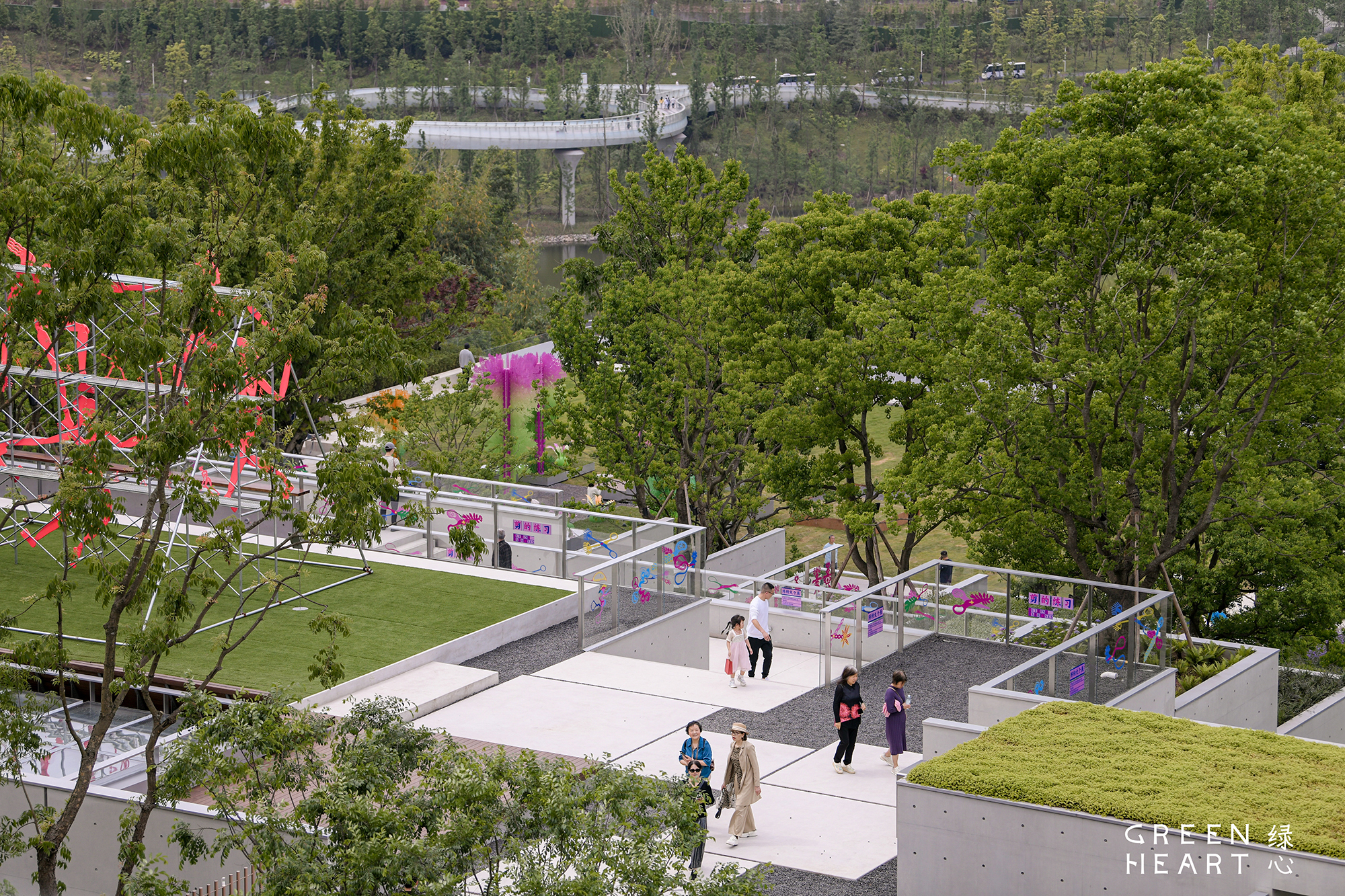 Rooftop terraces with glass balustrades, green roofs and walkers on pale paving, with a curving elevated bridge beyond the trees