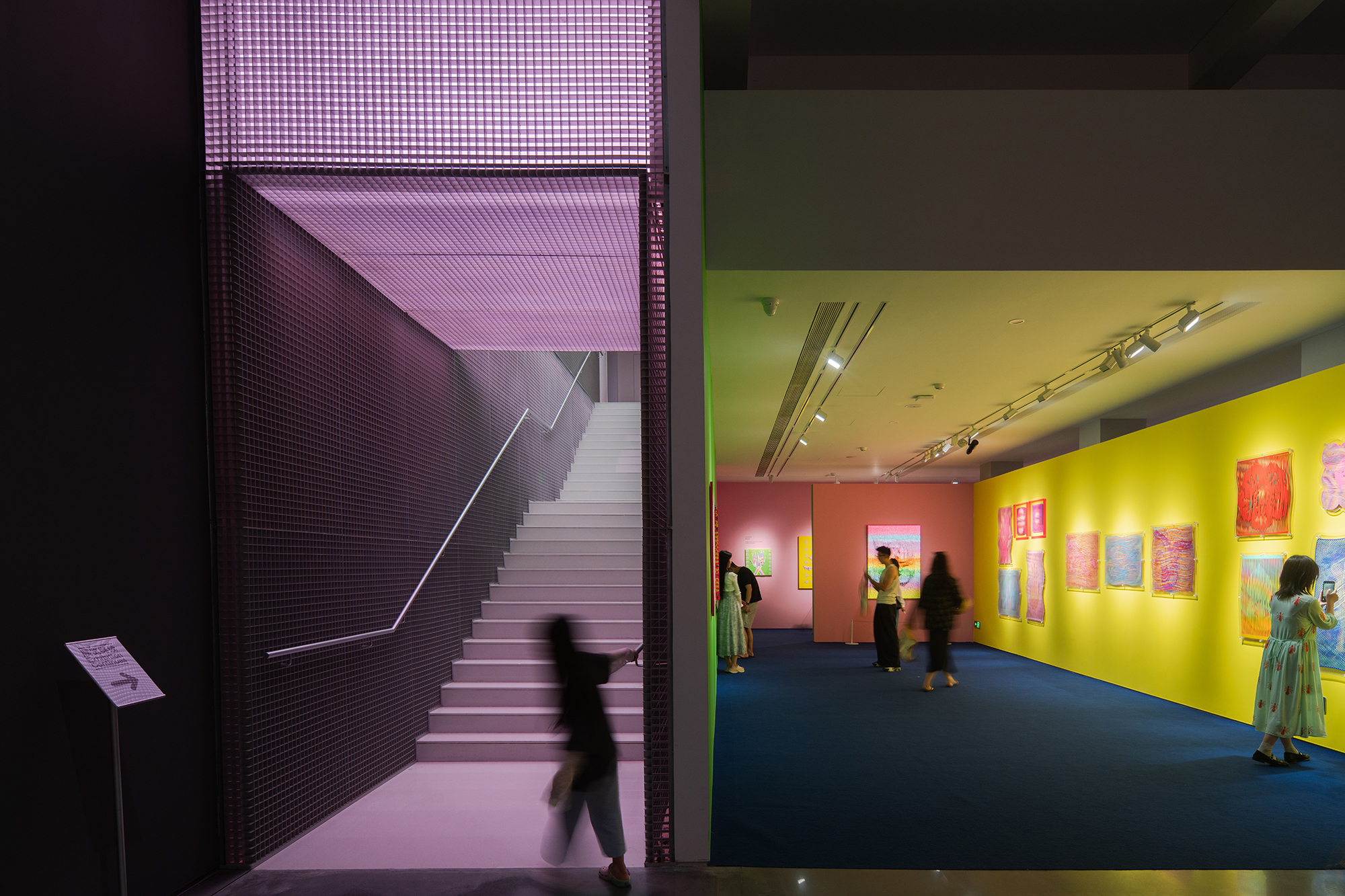 Gallery interior with a purple mesh-lined staircase beside a yellow wall of colorful paintings, with visitors moving through