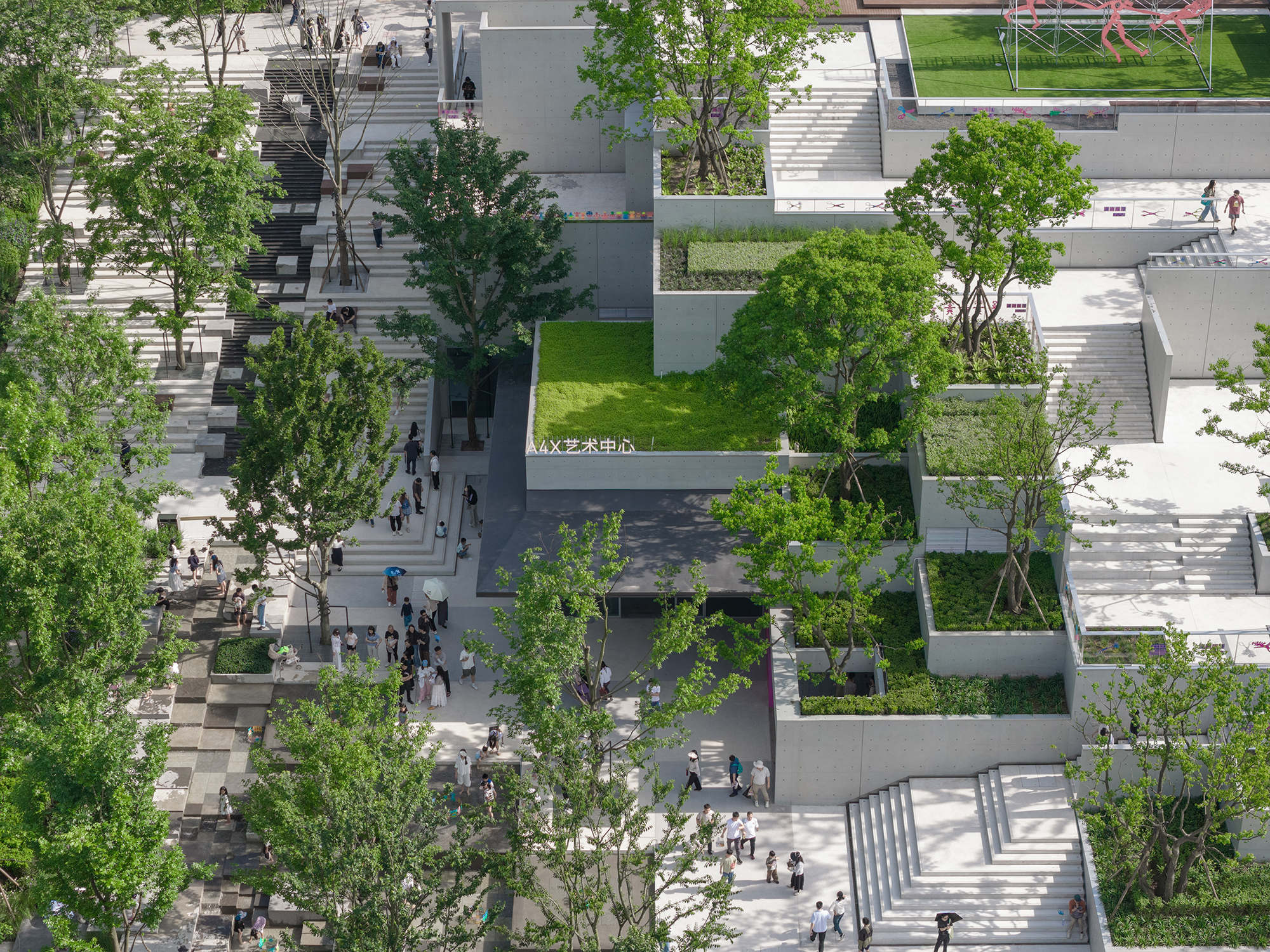 Elevated view of stepped concrete terraces with grass beds, young trees and visitors climbing wide white stairs on a bright day