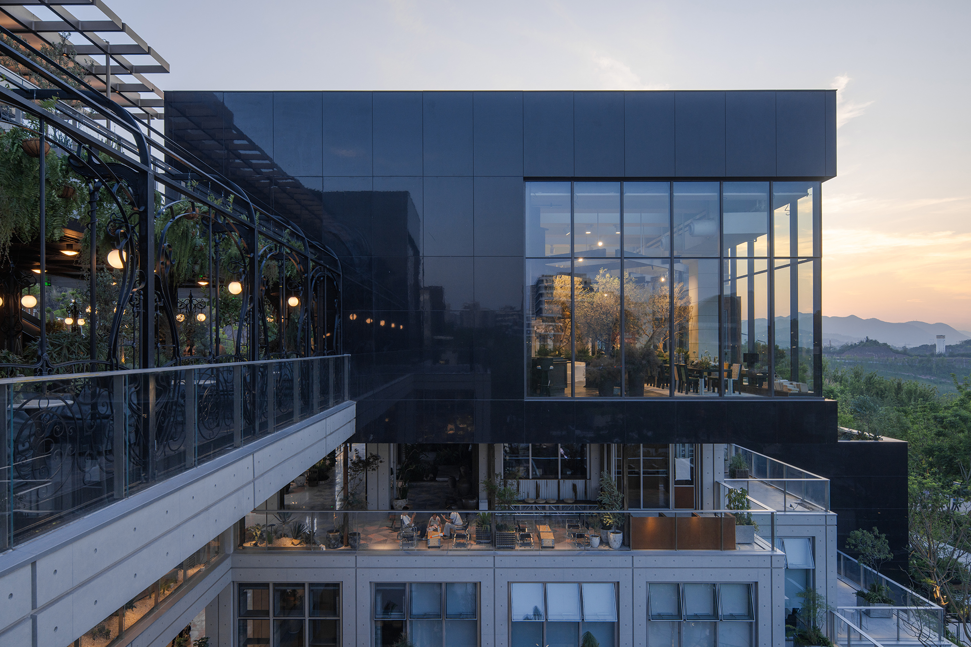 Dark glass-clad volume above a concrete base with an ornate iron-framed hanging garden terrace at dusk, hills glowing on the horizon