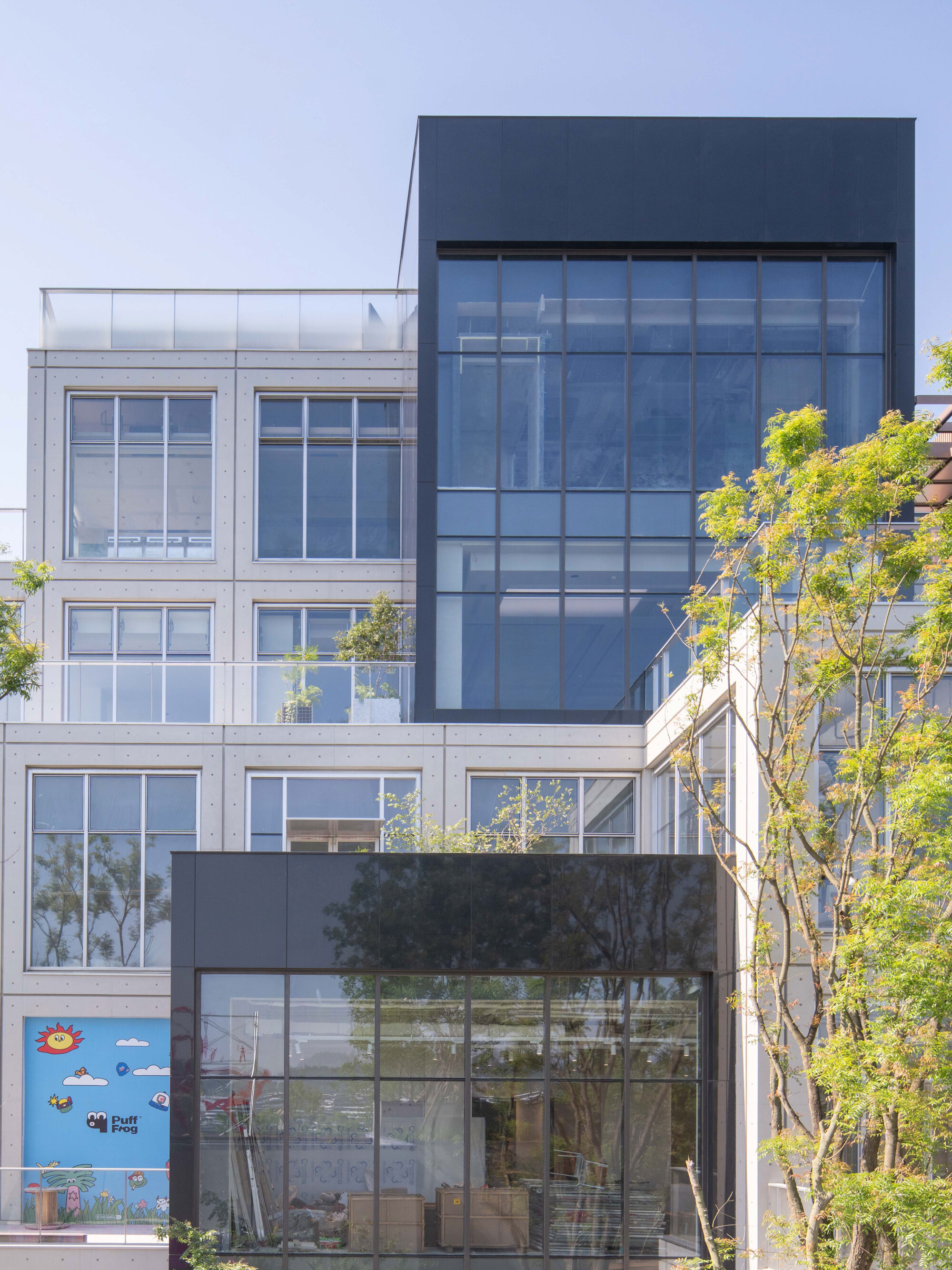 Facade of pale concrete panels and a black-framed glass box, with leafy branches in the foreground under a clear sky