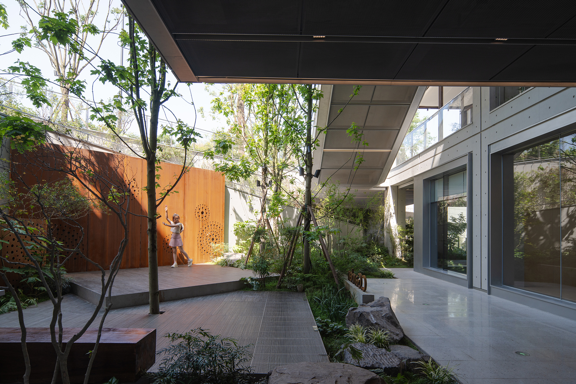 Garden courtyard with a perforated weathering steel screen, timber deck and young trees, a girl dancing in daylight