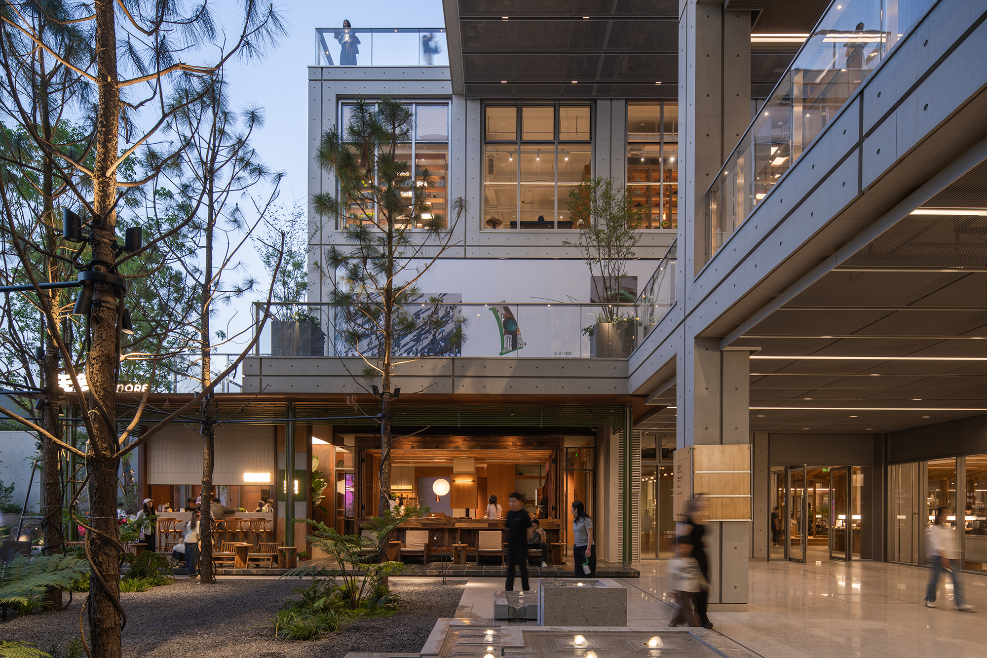 Courtyard with pines and gravel in front of a glazed cafe, stacked concrete floors above and blurred walkers at dusk